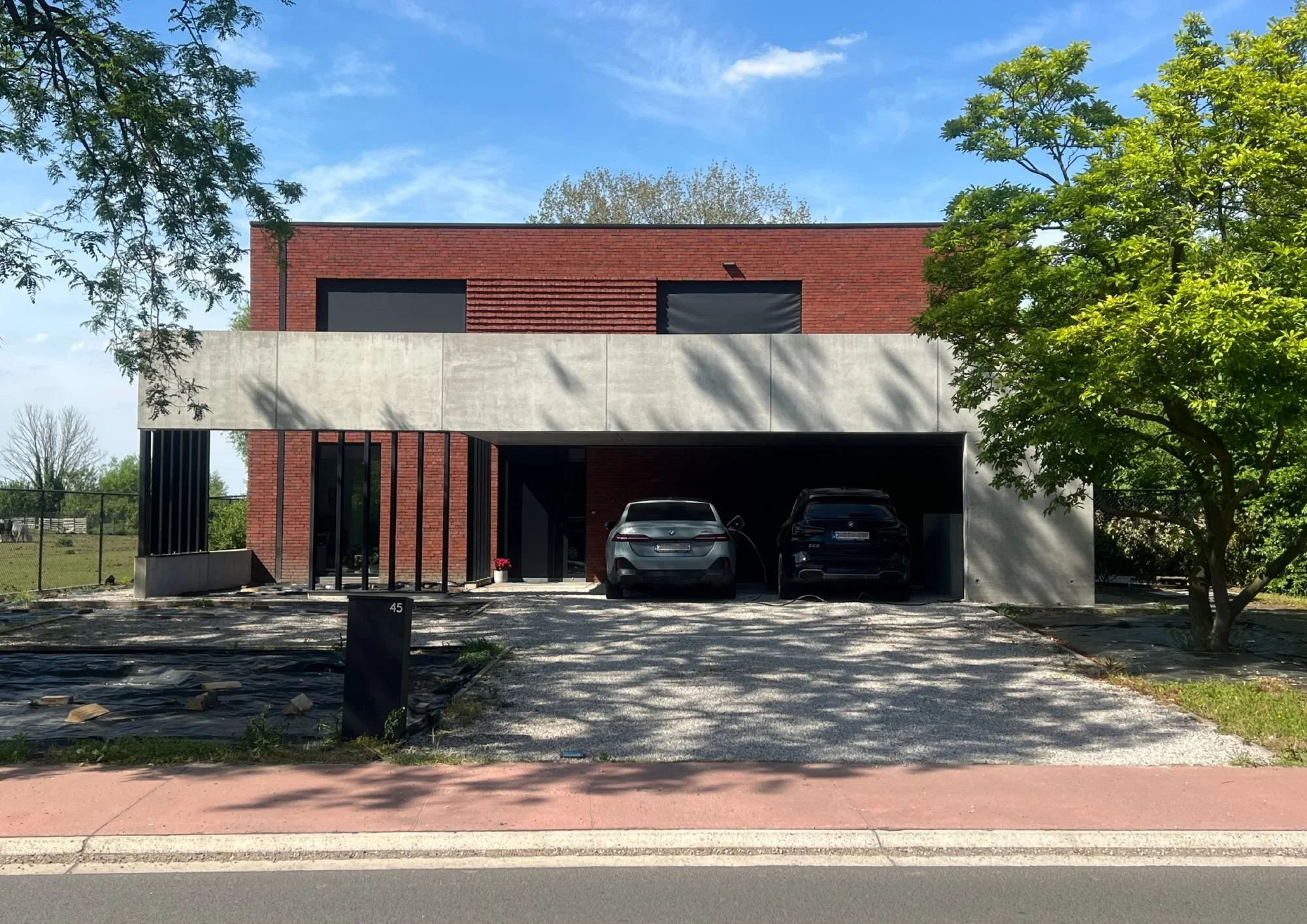 Modern building with brick and concrete exterior, parking area with two cars, surrounded by trees under a blue sky with clouds.