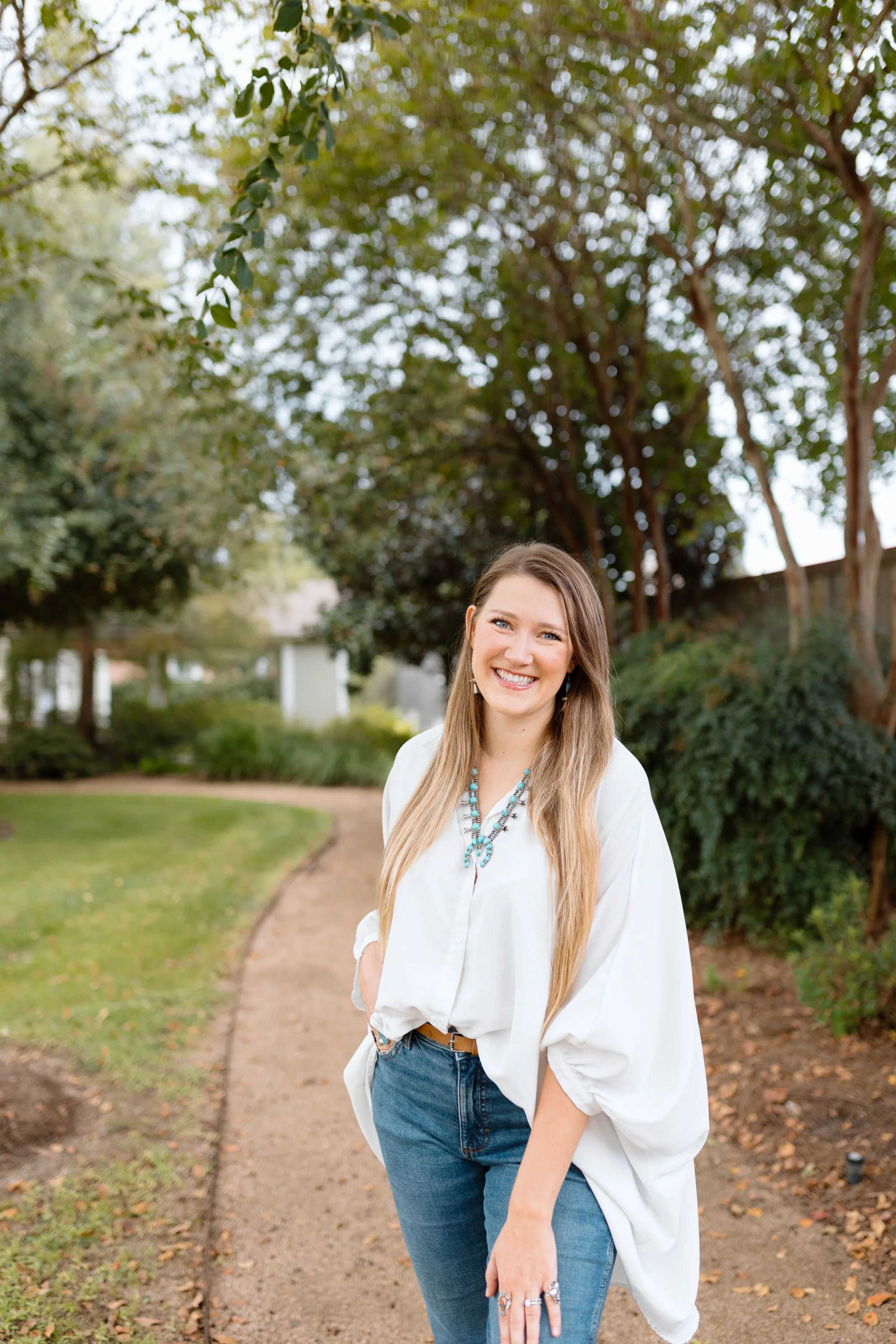A woman with long blonde hair, wearing a white blouse, blue jeans, and turquoise jewelry, smiling outdoors on a dirt path with green trees and bushes in the background.