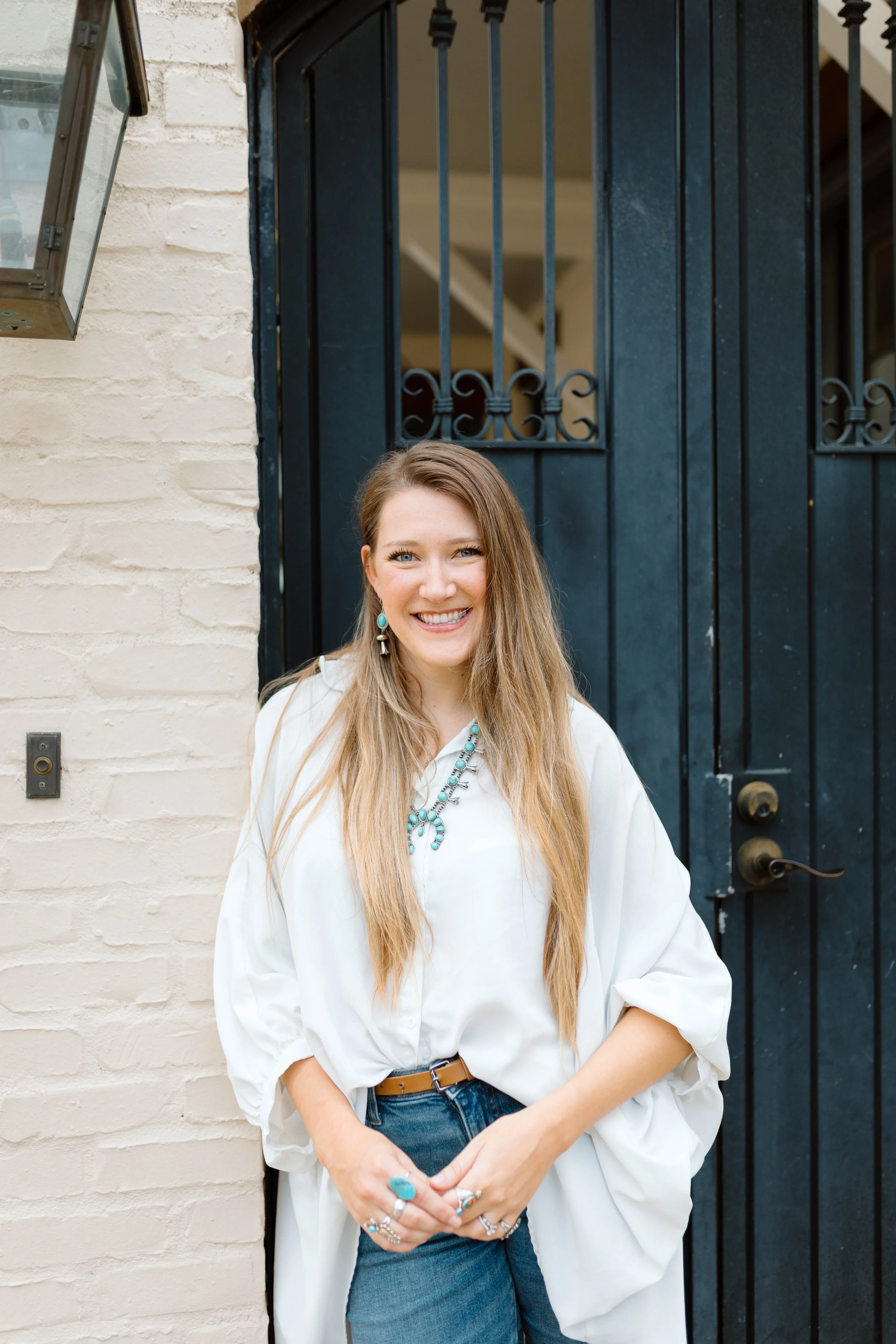 A smiling woman with long blonde hair wearing a white blouse, blue jeans, and turquoise jewelry standing in front of a black door with iron bars, next to a cream-colored brick wall.