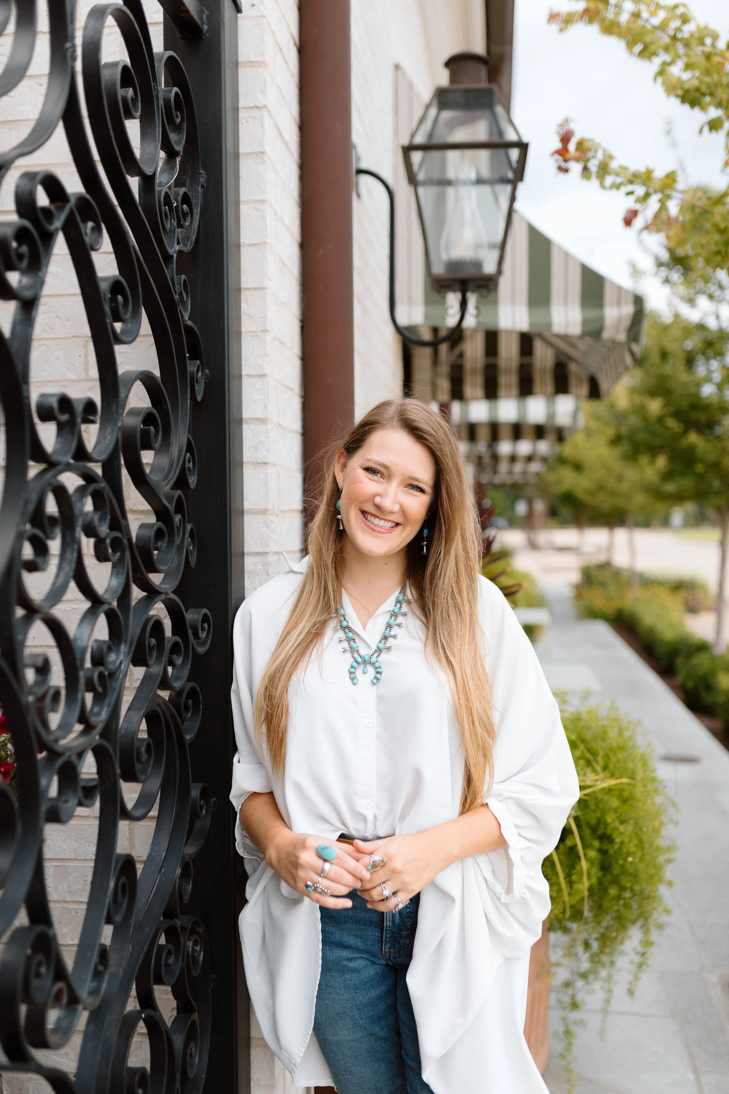 A smiling woman with long blonde hair stands on a sidewalk beside a white brick wall with decorative black ironwork. She wears a white shirt, blue jeans, and turquoise jewelry. A lantern and striped awnings are visible in the background, along with trees and greenery.
