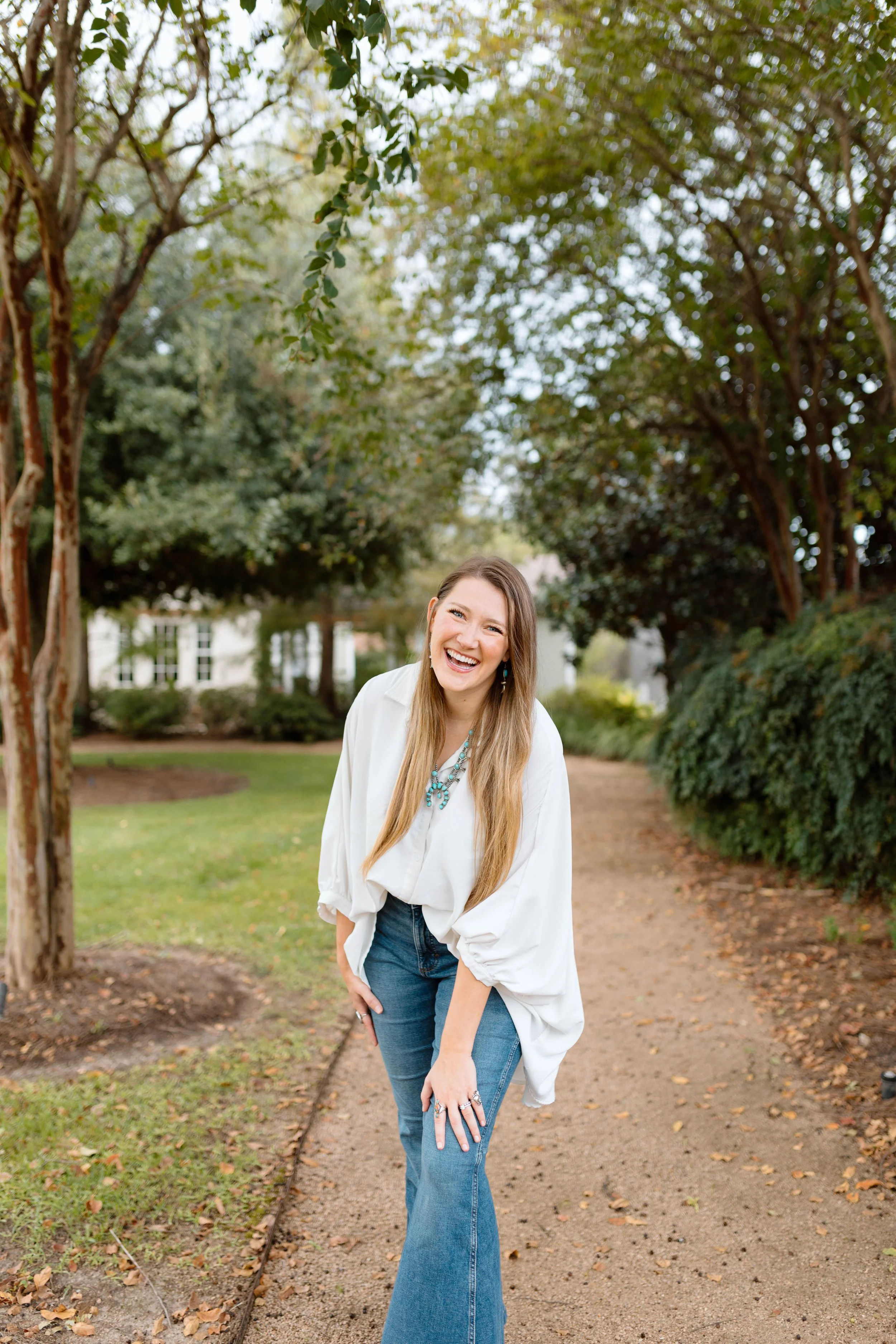 A woman with long hair smiling and laughing while standing on a dirt pathway in a park, wearing a white blouse, blue jeans, and turquoise jewelry, surrounded by green trees.