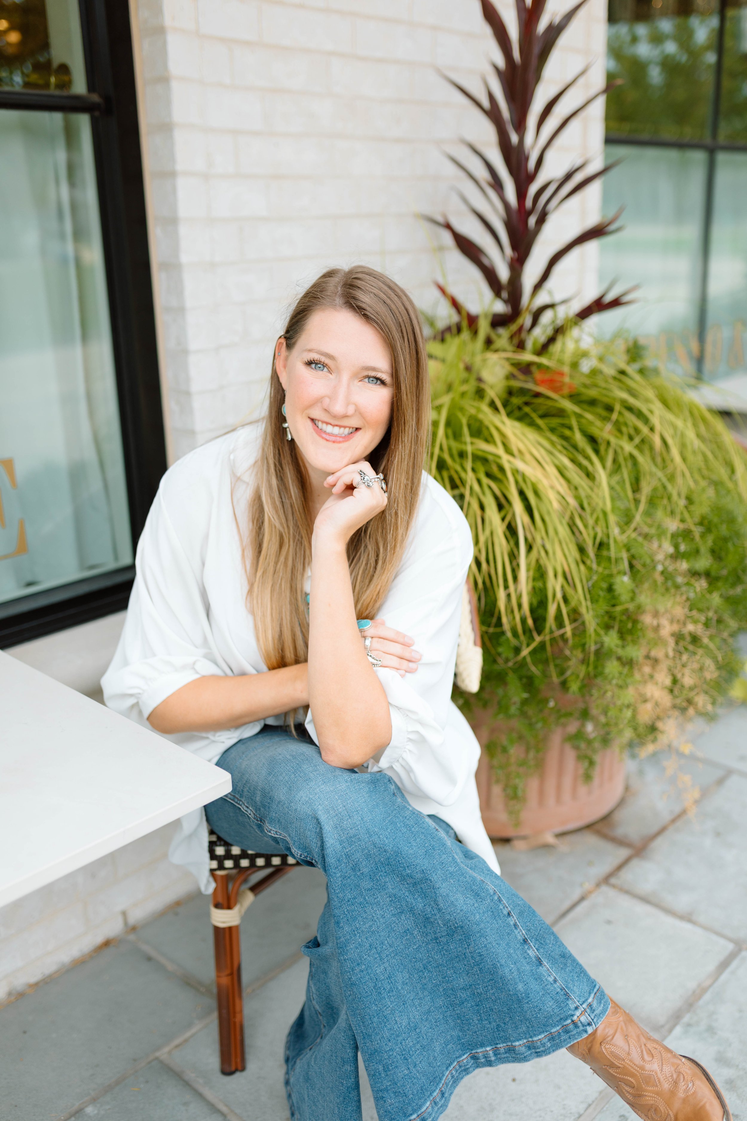 A smiling woman with long hair sitting outdoors near a large potted plant, wearing a white shirt, blue jeans, and cowboy boots.