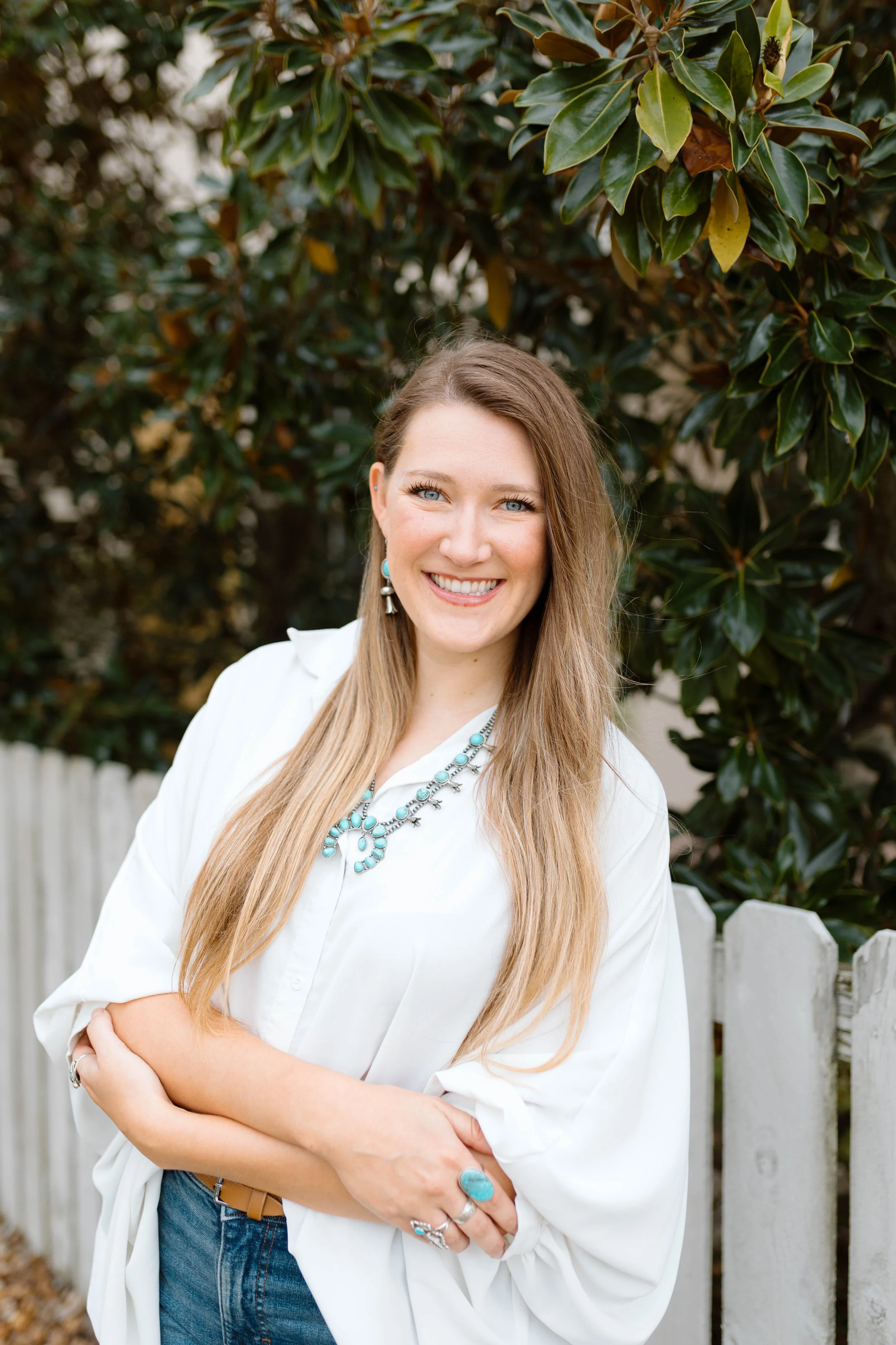 A smiling woman with long blonde hair, wearing a white shirt, blue jewelry, and a brown belt, standing outdoors in front of greenery and a white fence.