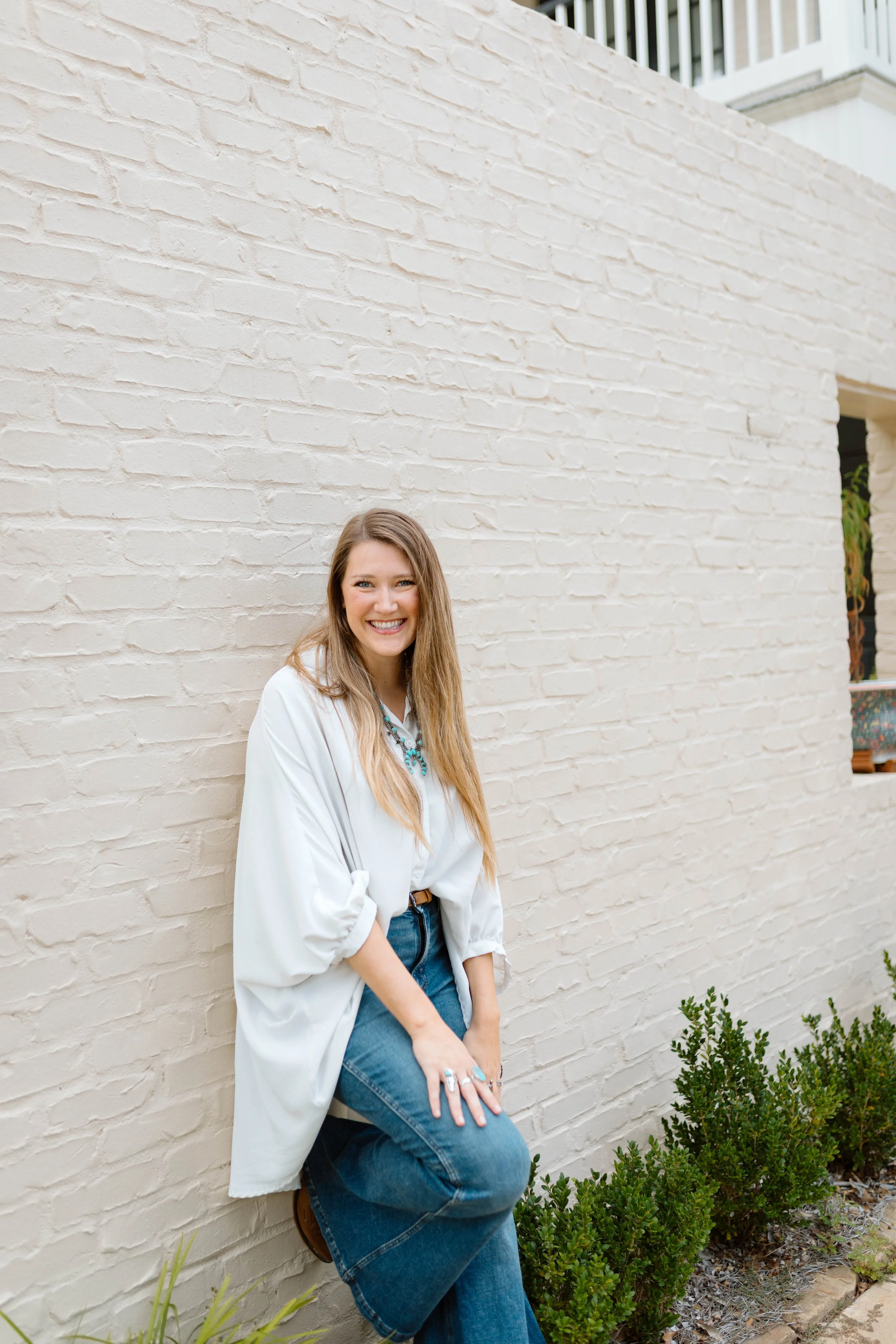 A young woman with long blonde hair smiling, leaning against a white brick wall outside, wearing a white blouse, blue jeans, and turquoise jewelry.