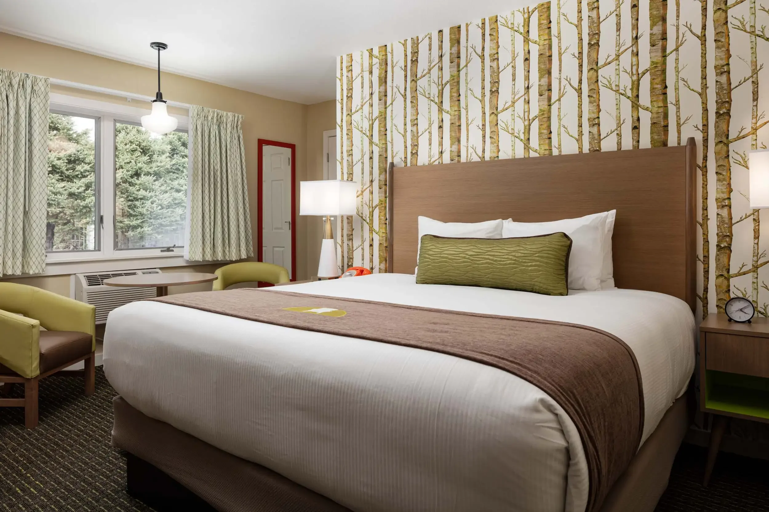A hotel room with a large bed, green decorative pillow, nightstand with clock, and a wallpaper featuring tree branches. There is a window with patterned curtains and a view of trees outside.