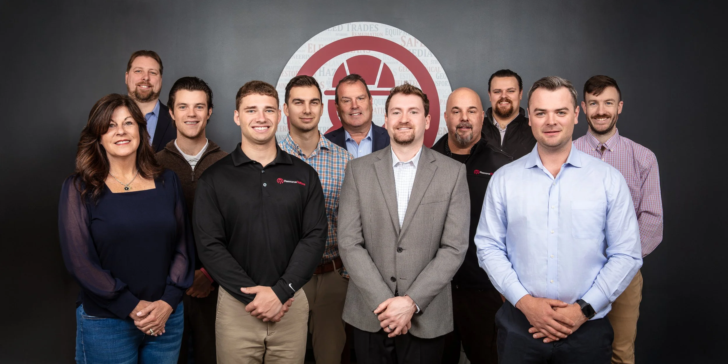 Group of eleven people standing together in front of a gray wall with a red and white logo. The group includes both men and women, all dressed in business casual or professional attire, smiling and posing for the photo.