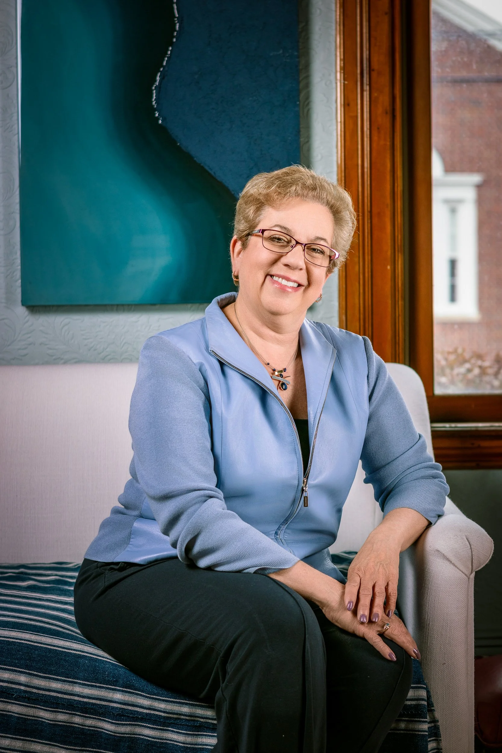 A smiling woman with short gray hair and glasses, sitting on a sofa with a striped cushion, in front of a window and a blue abstract painting.