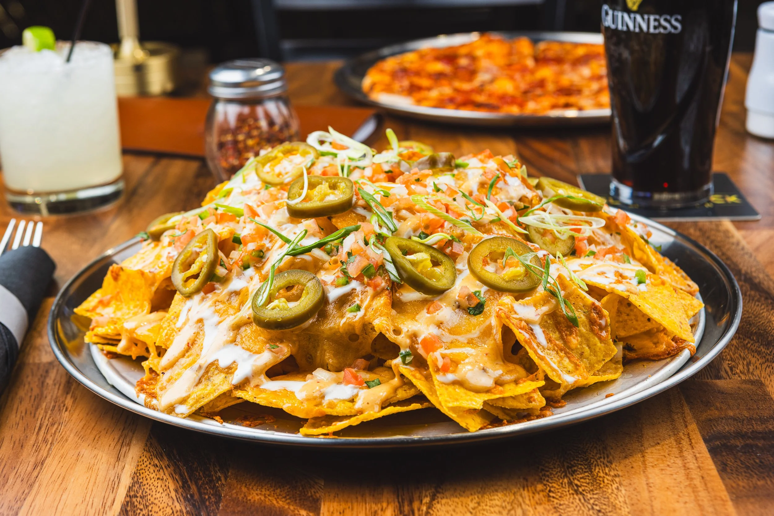 Plate of nachos topped with jalapeños, sour cream, diced tomatoes, green onions, and melted cheese on a wooden table with drinks and pizza in the background.