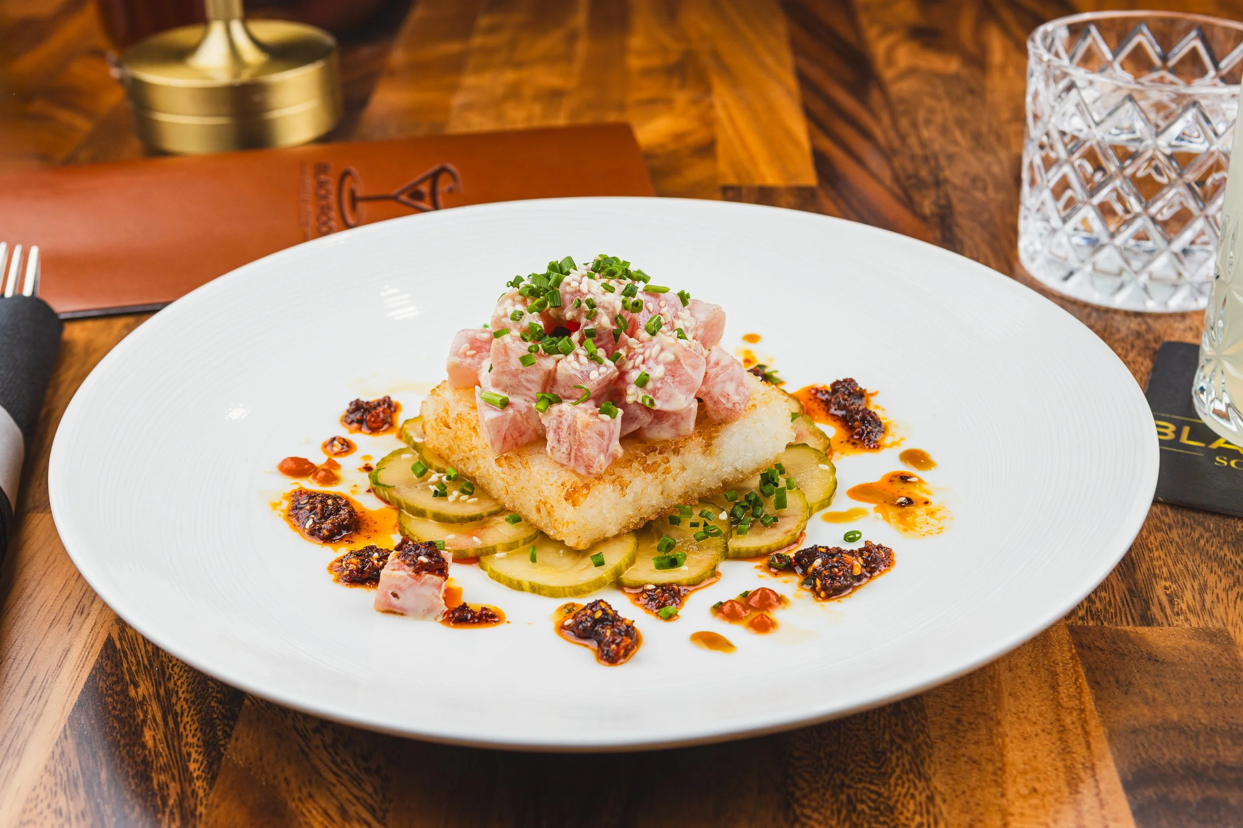 A plated dish with sliced cucumbers, a piece of fried fish, and chopped raw fish with herbs, garnished with chives and spicy sauce drizzles on a white plate on a wooden table.
