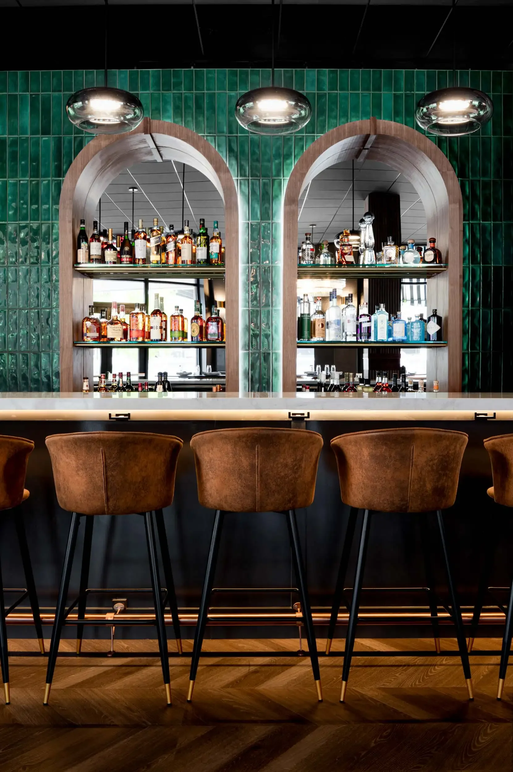 A bar with a green tiled wall, wooden arches, and shelves filled with various bottles of liquor. There are four brown bar stools with black legs and gold accents in front of the bar. The flooring is a dark wooden pattern.