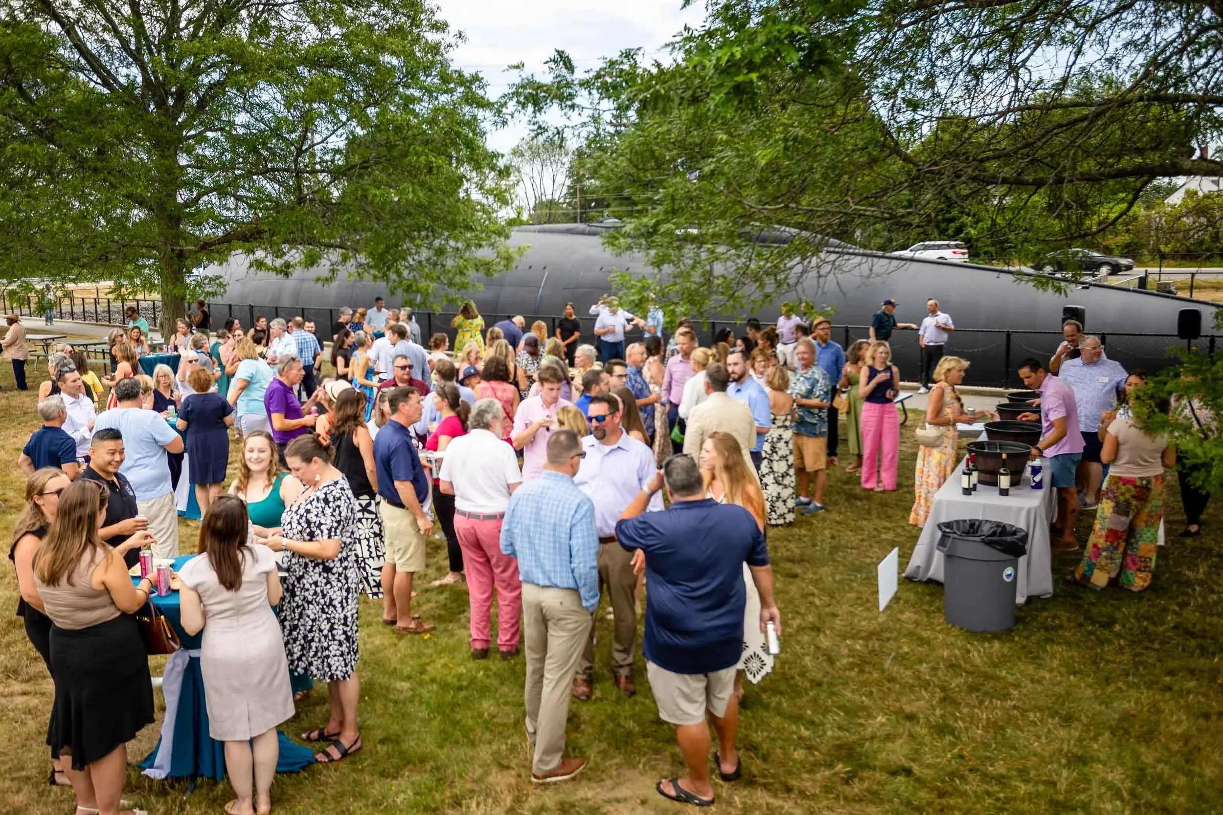 A large gathering of people at an outdoor event, standing around and socializing on a grassy area under trees, with food and drink tables, and a black submarine display in the background.
