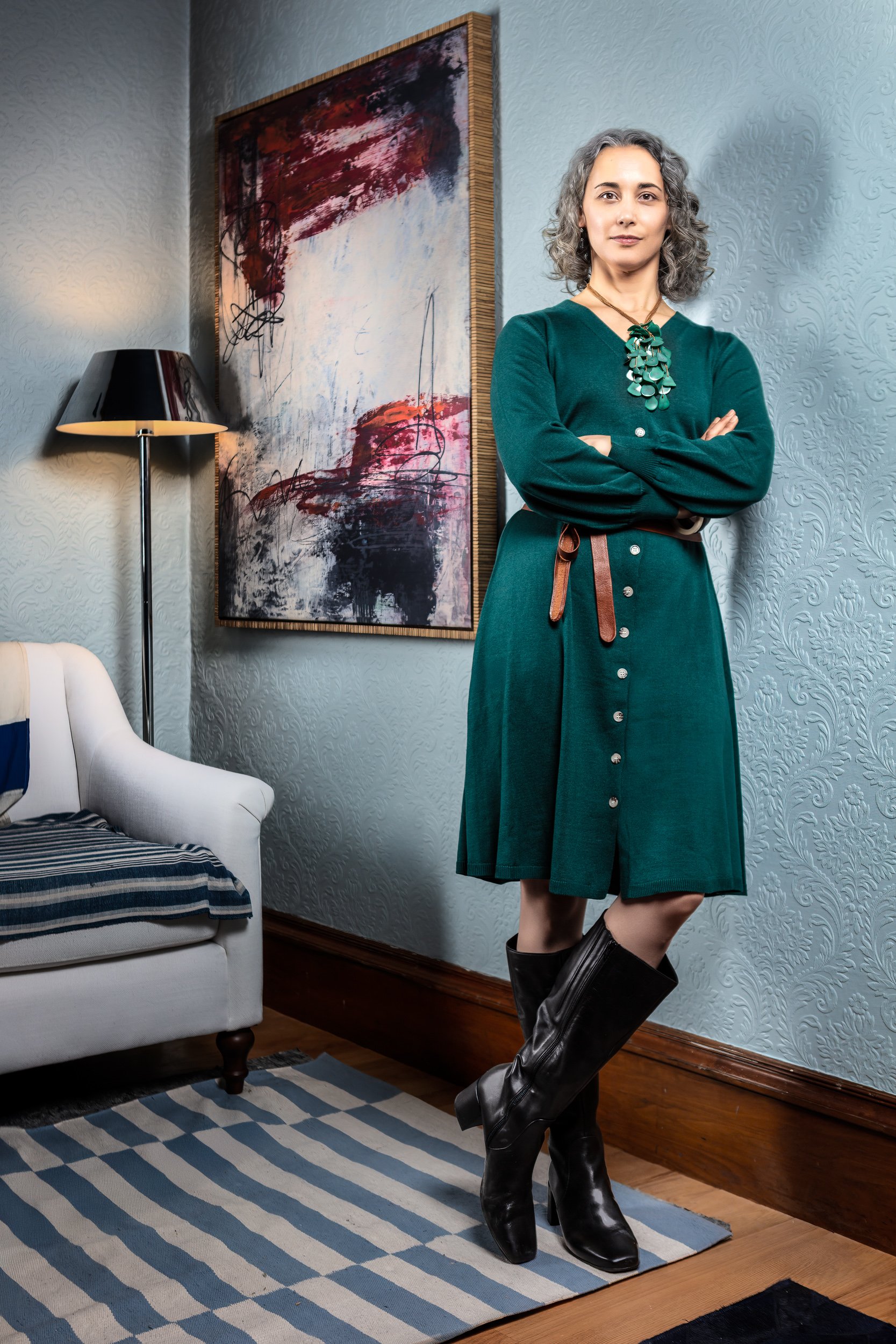 A woman with curly gray hair in a teal dress and black knee-high boots standing in a room with light blue textured wallpaper, a black lamp, and an abstract painting on the wall.