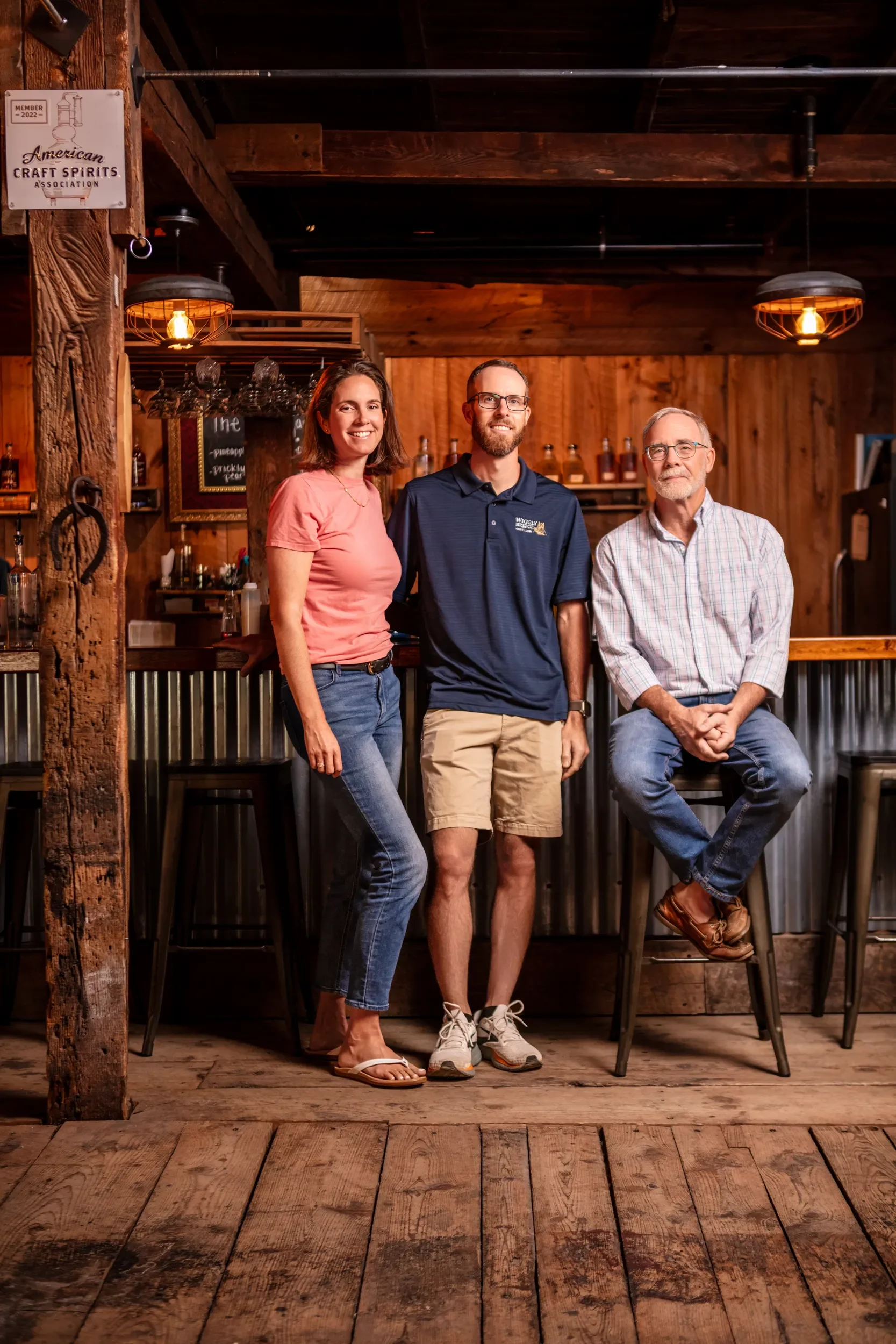 Three people standing in a rustic bar with wood-paneled walls and a corrugated metal bar front. The woman on the left is wearing a pink t-shirt and jeans, smiling. The man in the middle is wearing glasses, a navy polo shirt, and khaki shorts, smiling