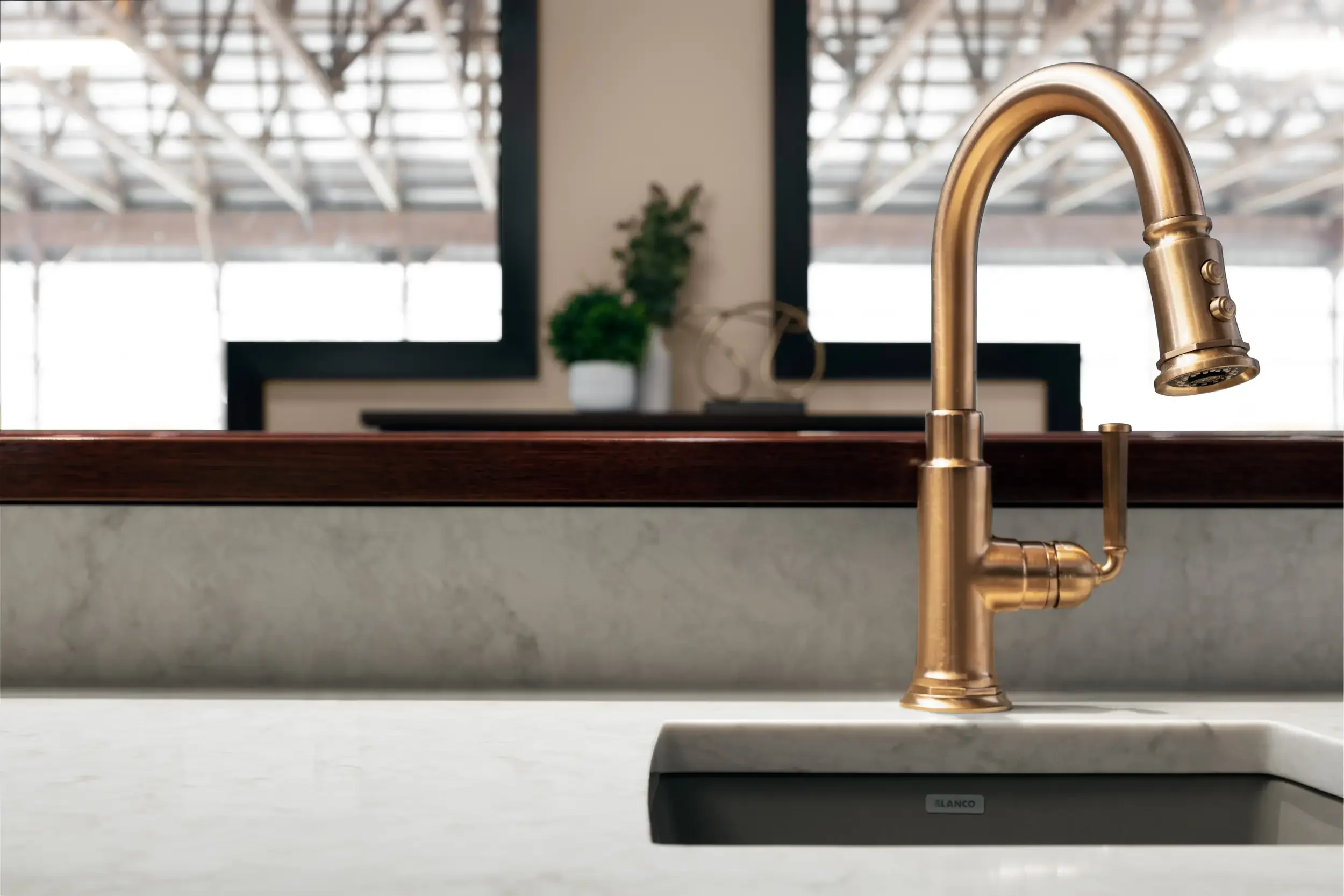 Close-up of a modern kitchen faucet with a gold finish installed on a white countertop, with a blurred background showing a window and a green plant.