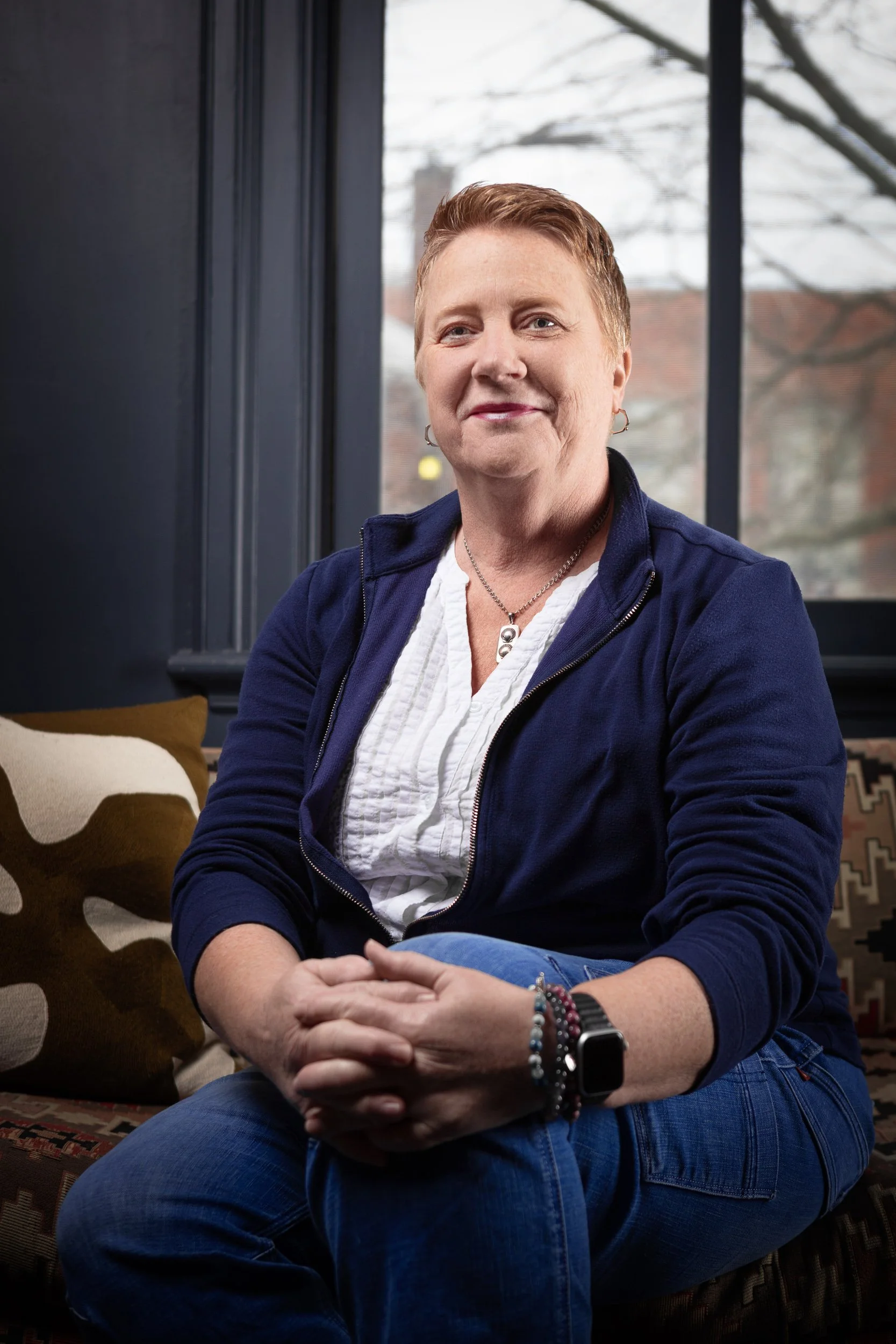 A woman with short red hair and fair skin sitting on a patterned sofa in front of a window outdoors. She wears a navy blue zip-up jacket, a white blouse, blue jeans, and accessories including bracelets, earrings, a necklace, and a smartwatch. She is 