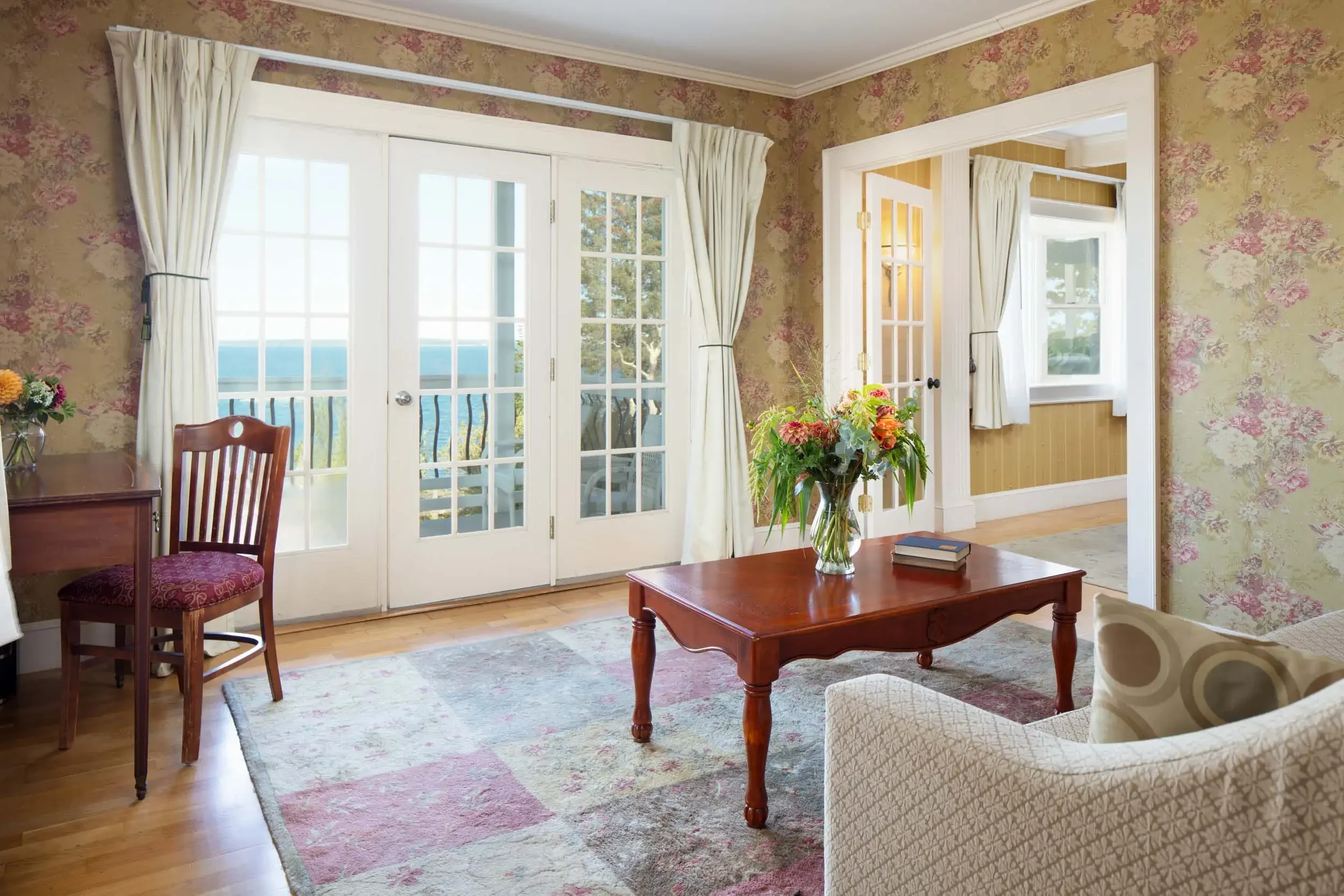 Living room with floral wallpaper, large glass door with white curtains, and a window with a view outside. There is a wooden table with a vase of flowers, a book, and part of an armchair visible. A chair with a cushioned seat and backrest is near the