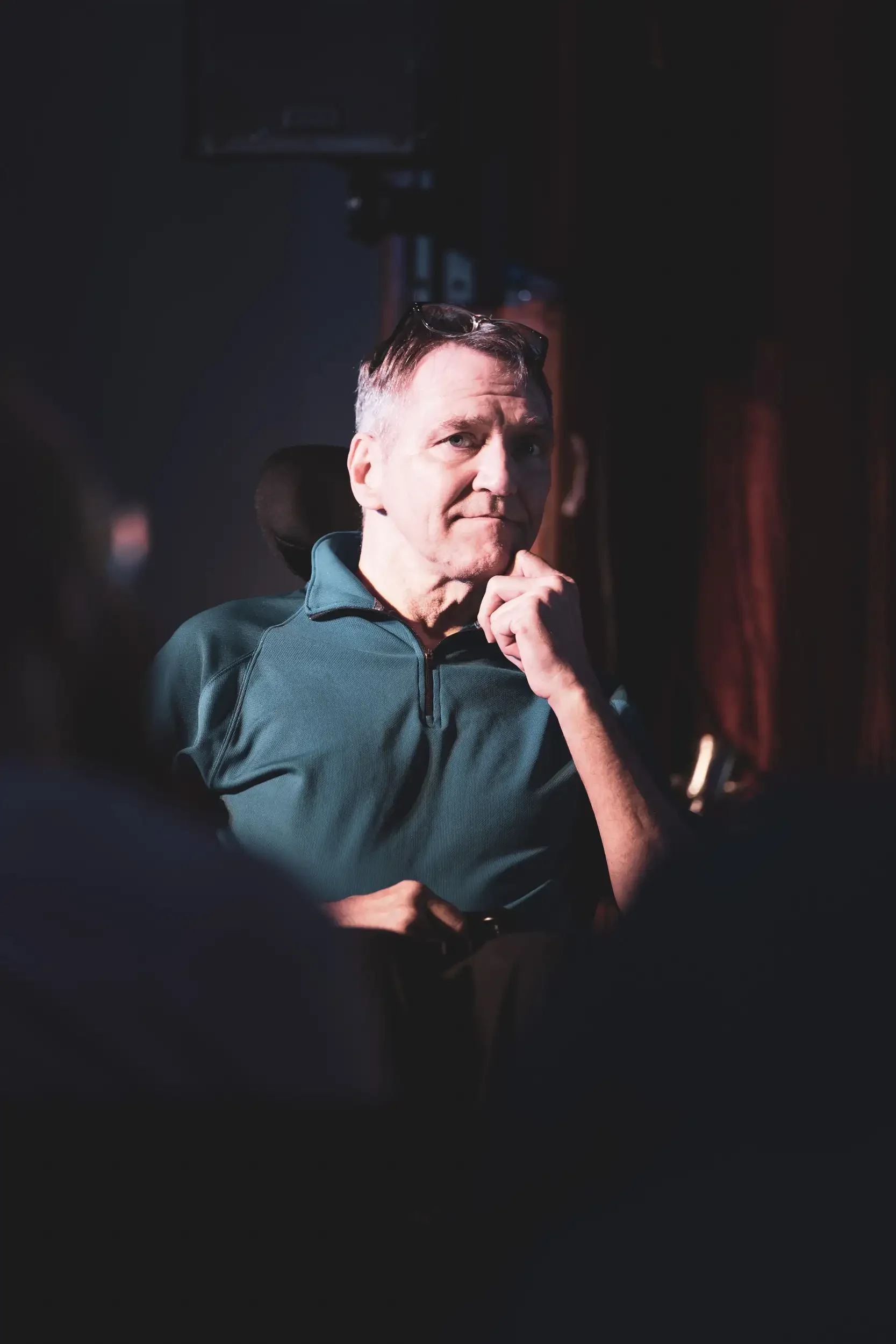 A man with short gray hair, blue eyes, and wearing a teal athletic jacket, sitting in a dark room with a serious expression, glasses resting on his head, pensive pose with hand near chin.