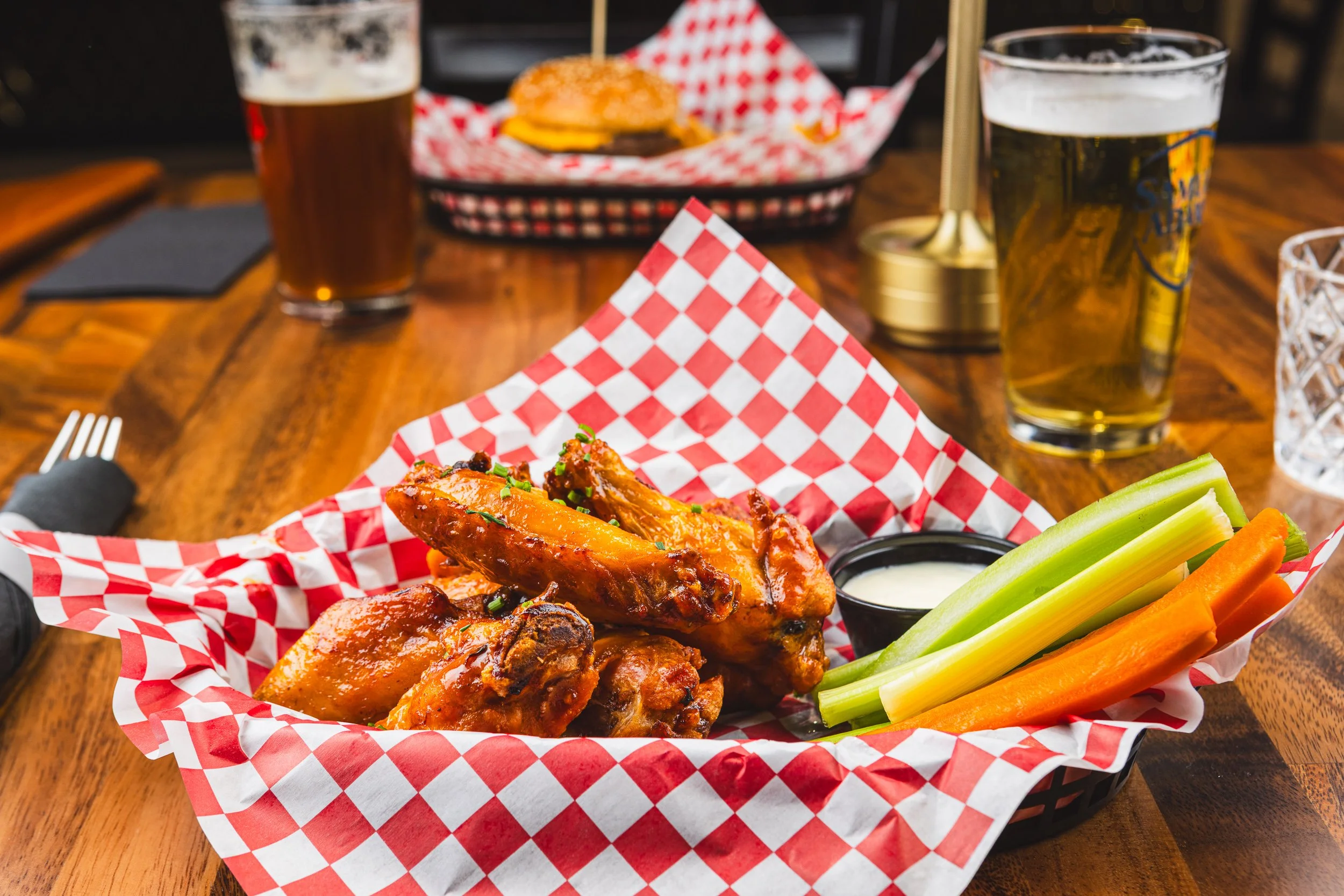 Basket of chicken wings with celery and carrot sticks, accompanied by dipping sauce, on a wooden table in a restaurant setting. In the background, there are two glasses of beer and a burger in a basket.