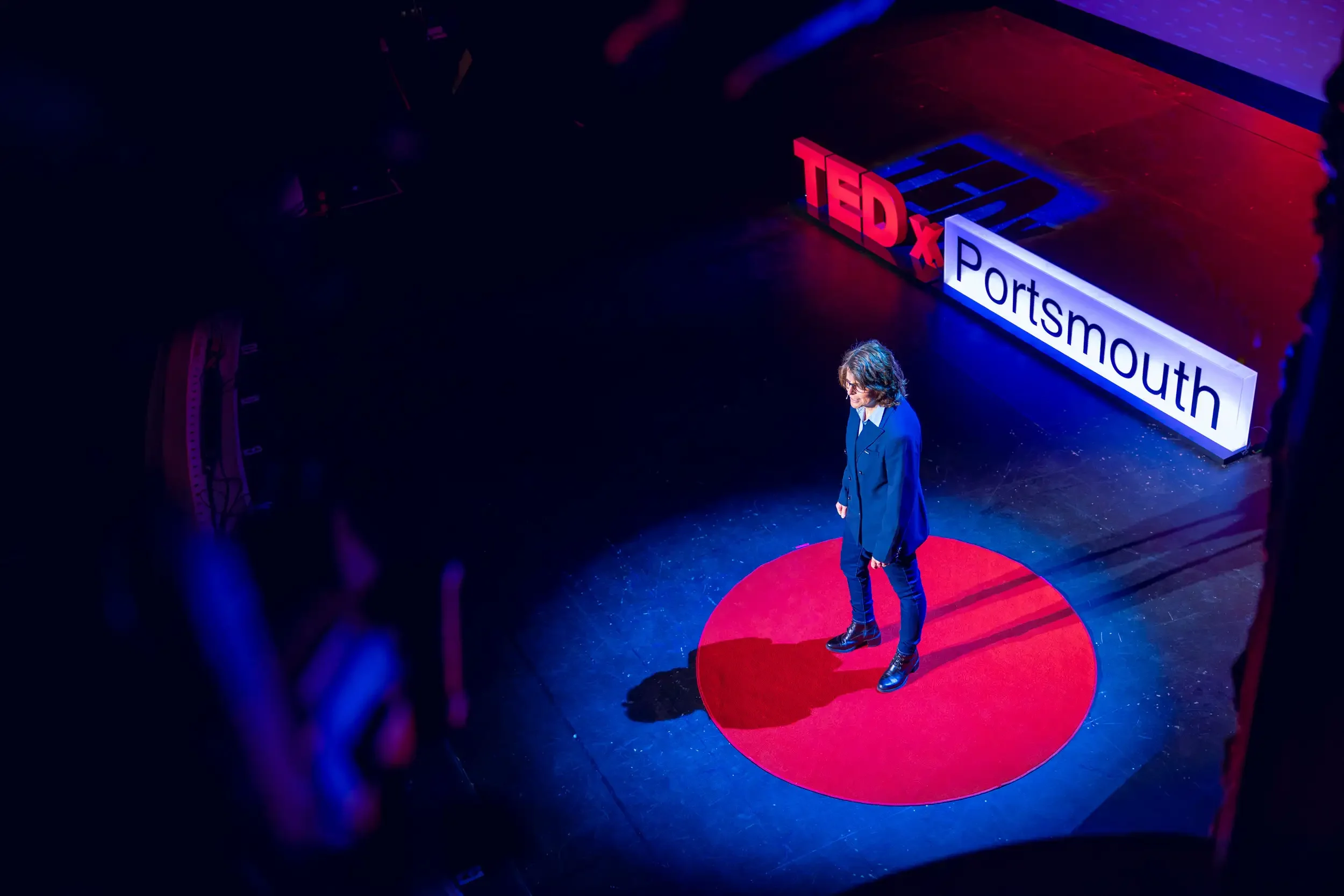 A woman stands on a red circular stage during a TEDx event at Sportsmouth. Signs with 'TEDx' and 'Sportsmouth' are visible behind her.
