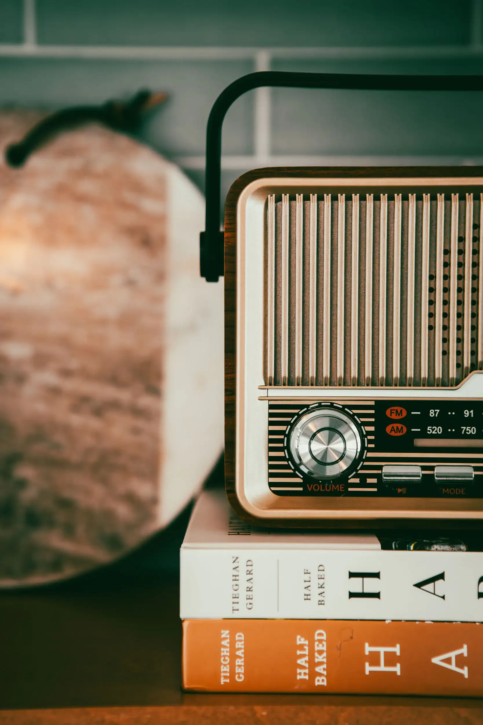 Close-up of a vintage radio placed on top of a Stack of books, including titles like 'Half Bakery' and 'The'. Part of a person's arm and a blurred background are also visible.