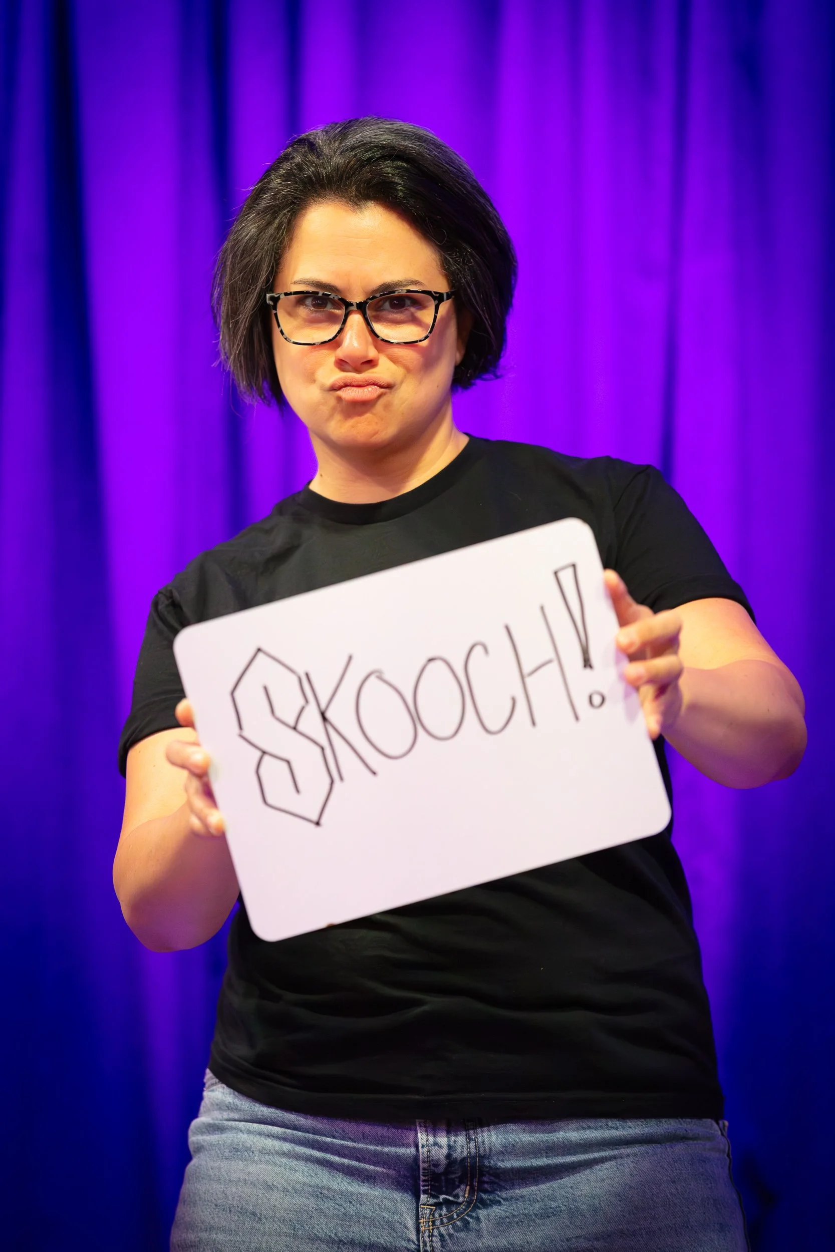 Woman with short dark hair and glasses holding a whiteboard that has '$KOCH!' written on it, standing in front of a purple curtain.