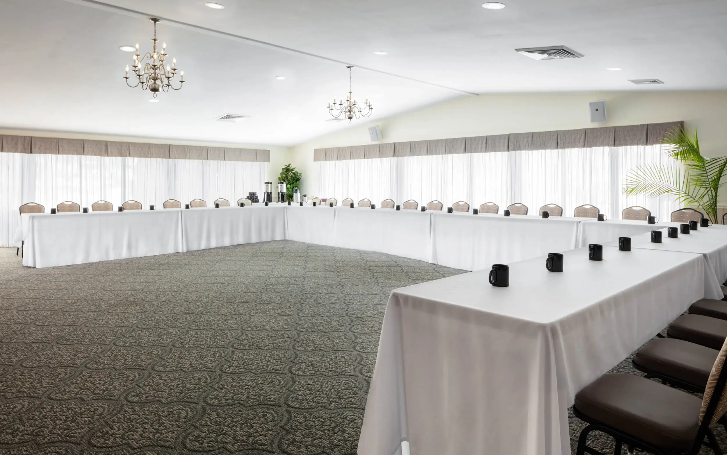 A large conference room with long tables arranged in a U-shape, covered with white tablecloths, with black coffee mugs, beige cushioned chairs, hanging chandeliers, and large windows with white curtains and valances, decorated with potted plants.