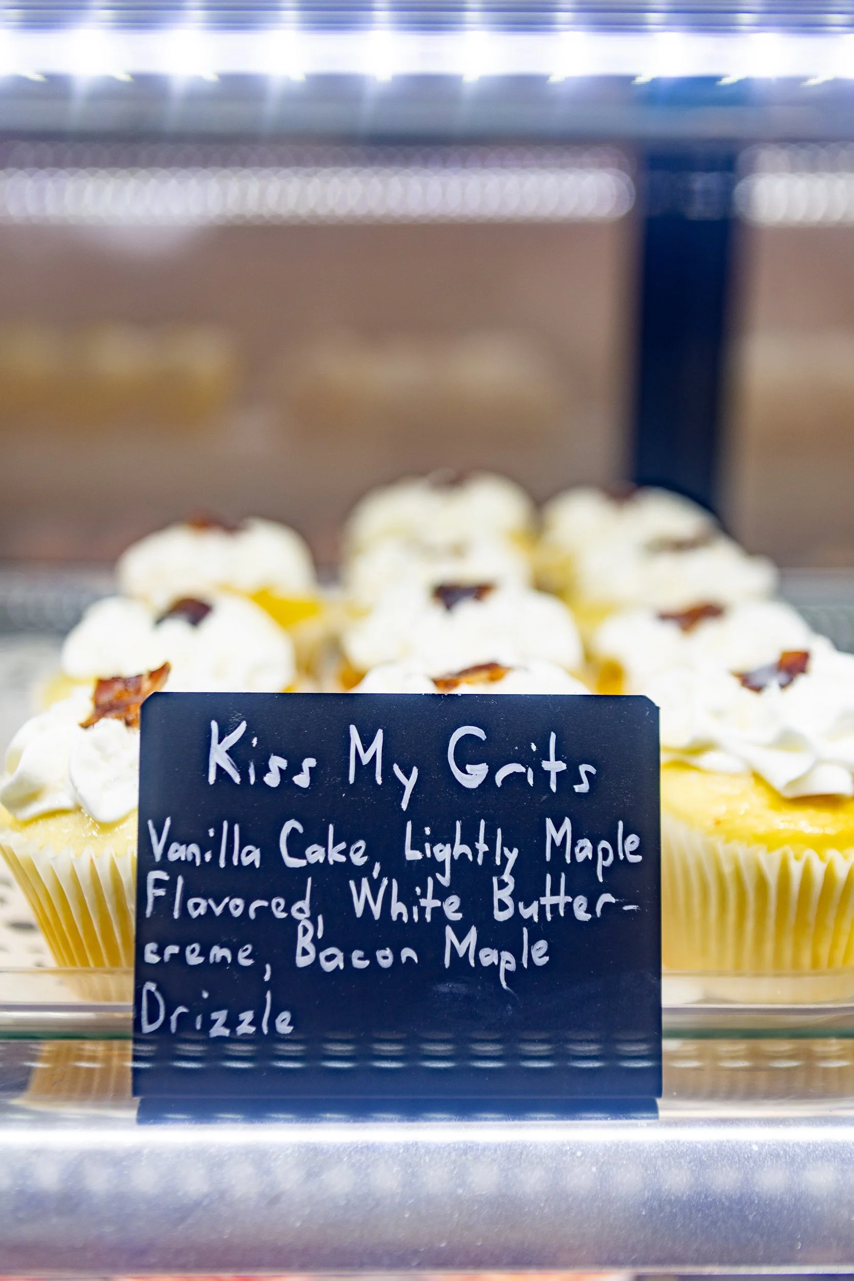 Display of vanilla cupcakes with white frosting and bacon bits on top, labeled "Kiss My Grits: Vanilla Cake, Lightly Maple Flavored White Buttercream, Bacon Maple Drizzle."