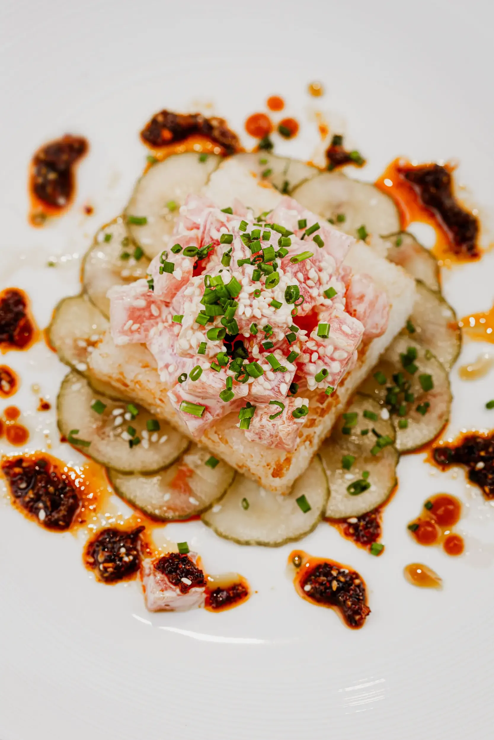 Close-up of a plated dish with sliced vegetables, a pink and white seafood salad garnished with chopped green onions, surrounded by drizzled sauces and chili paste.