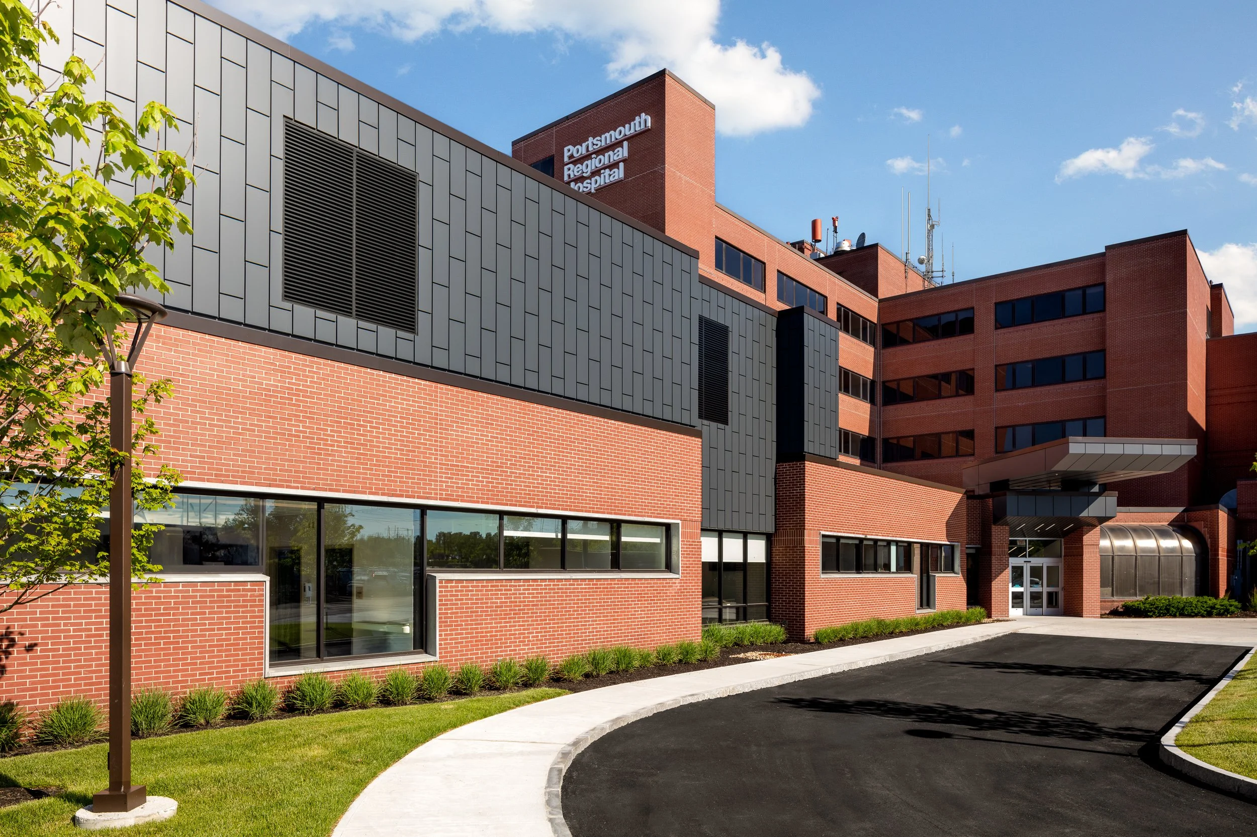 Exterior view of Portsmouth Regional Hospital, a multi-story brick and metal building with a curved driveway, green lawn, and a tree in the foreground under a blue sky with clouds.
