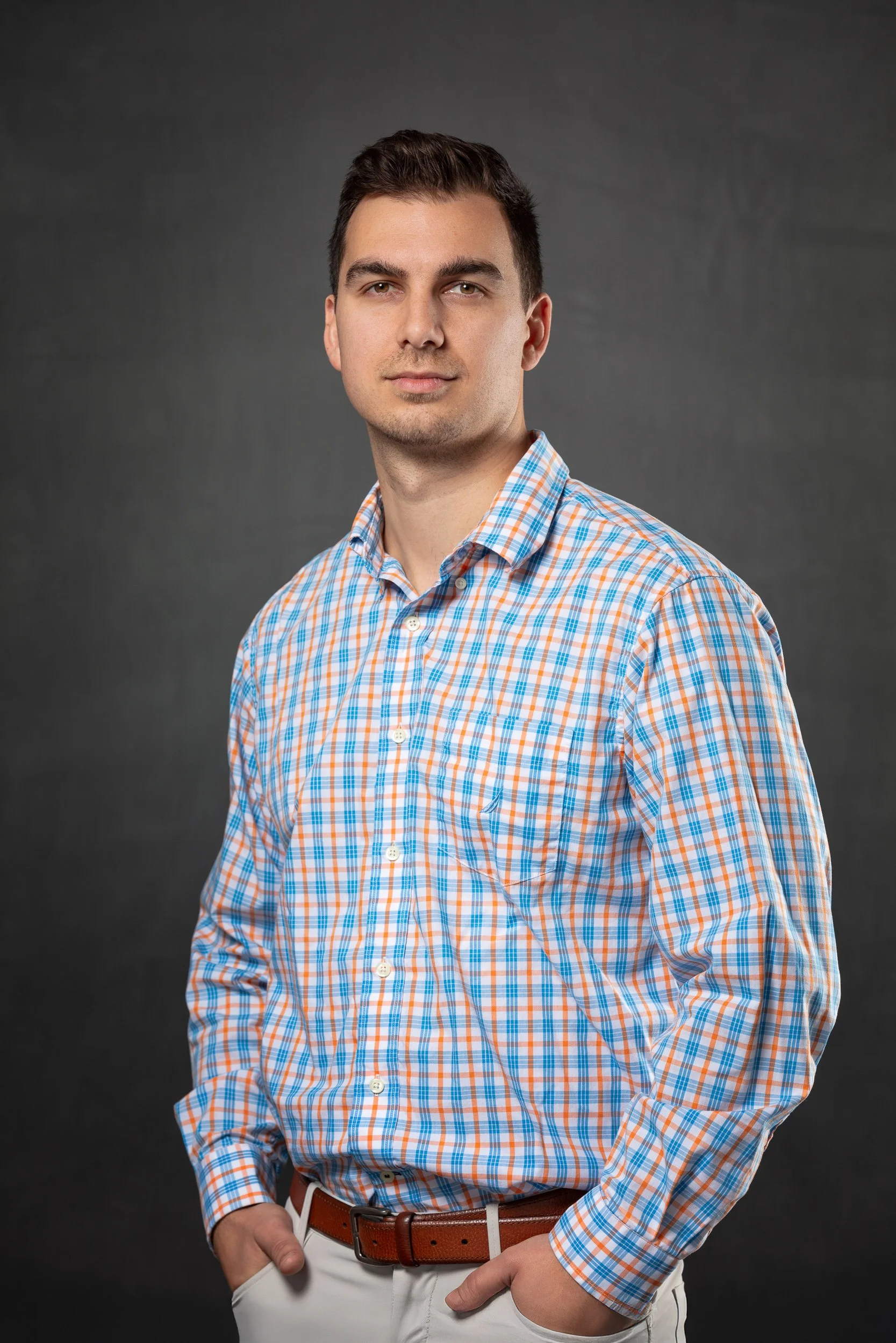 A young man with short dark hair and light skin standing against a dark gray background, wearing a button-down shirt with a orange, blue, and white plaid pattern and white pants, with his hands in his pockets.