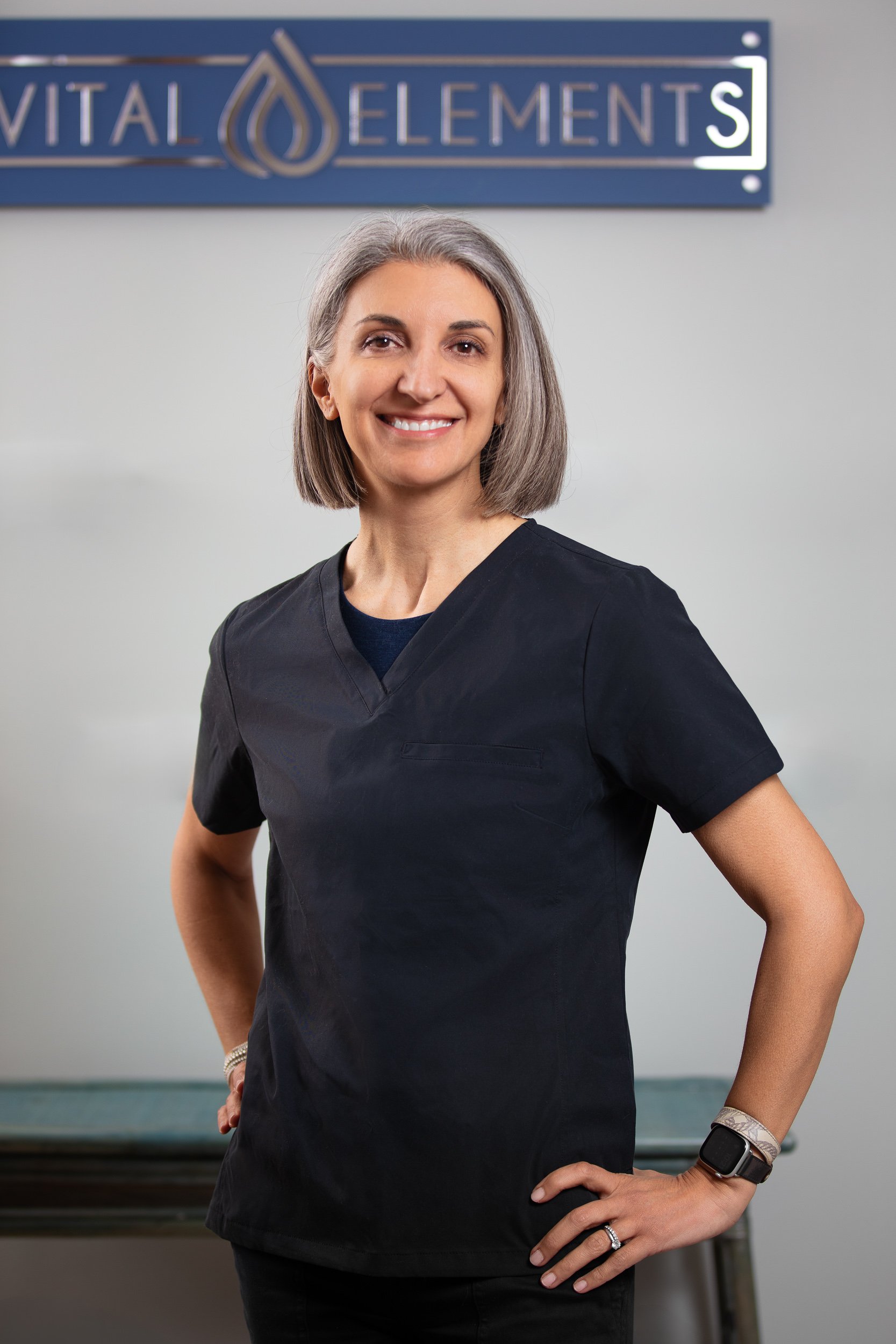 A smiling woman with gray hair and wearing black scrubs standing in front of a sign that reads 'VITAL ELEMENTS'.