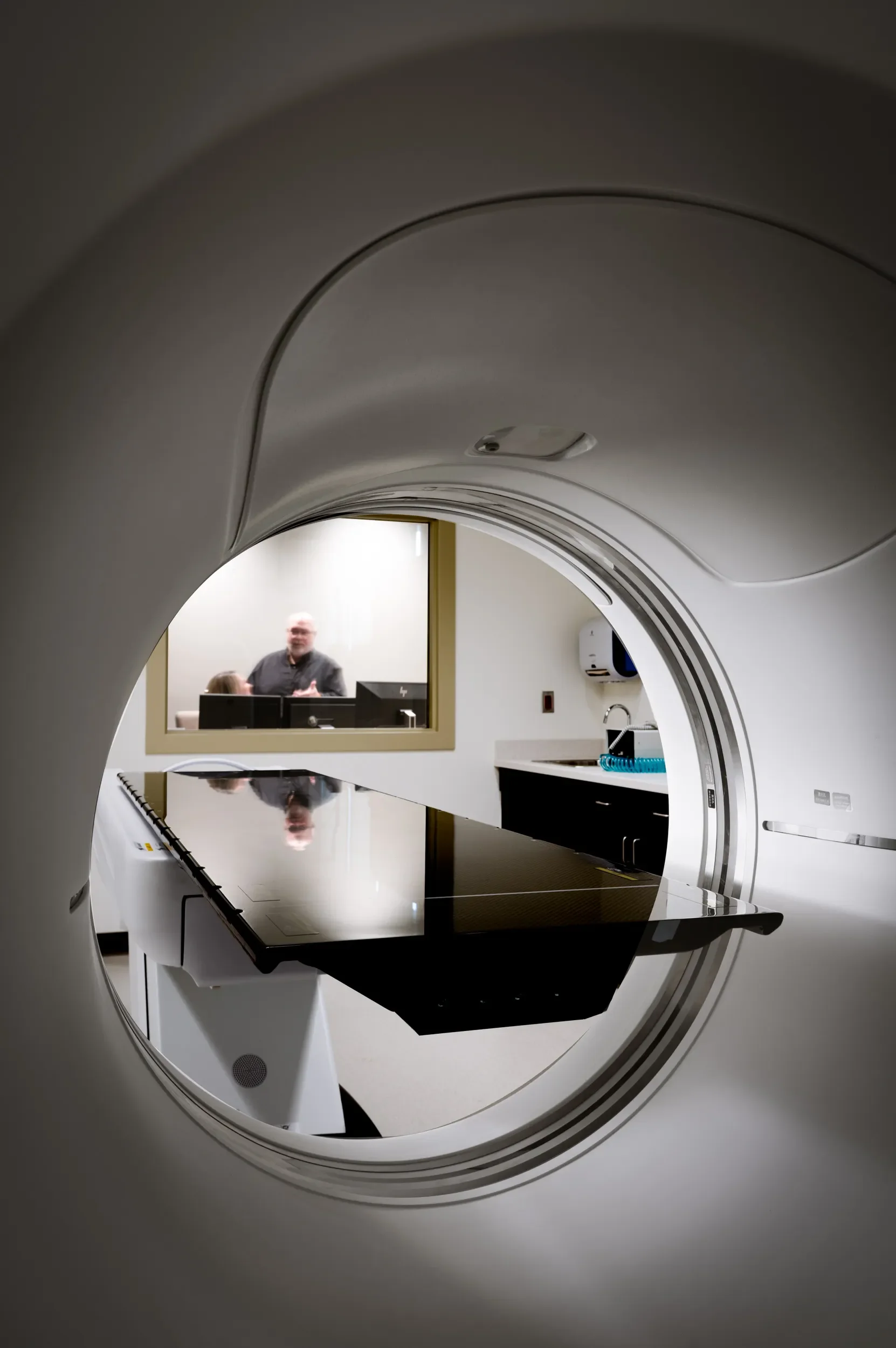 Interior view of a medical imaging room showing a CT scanner with a patient bed and a technician or doctor talking through a window.