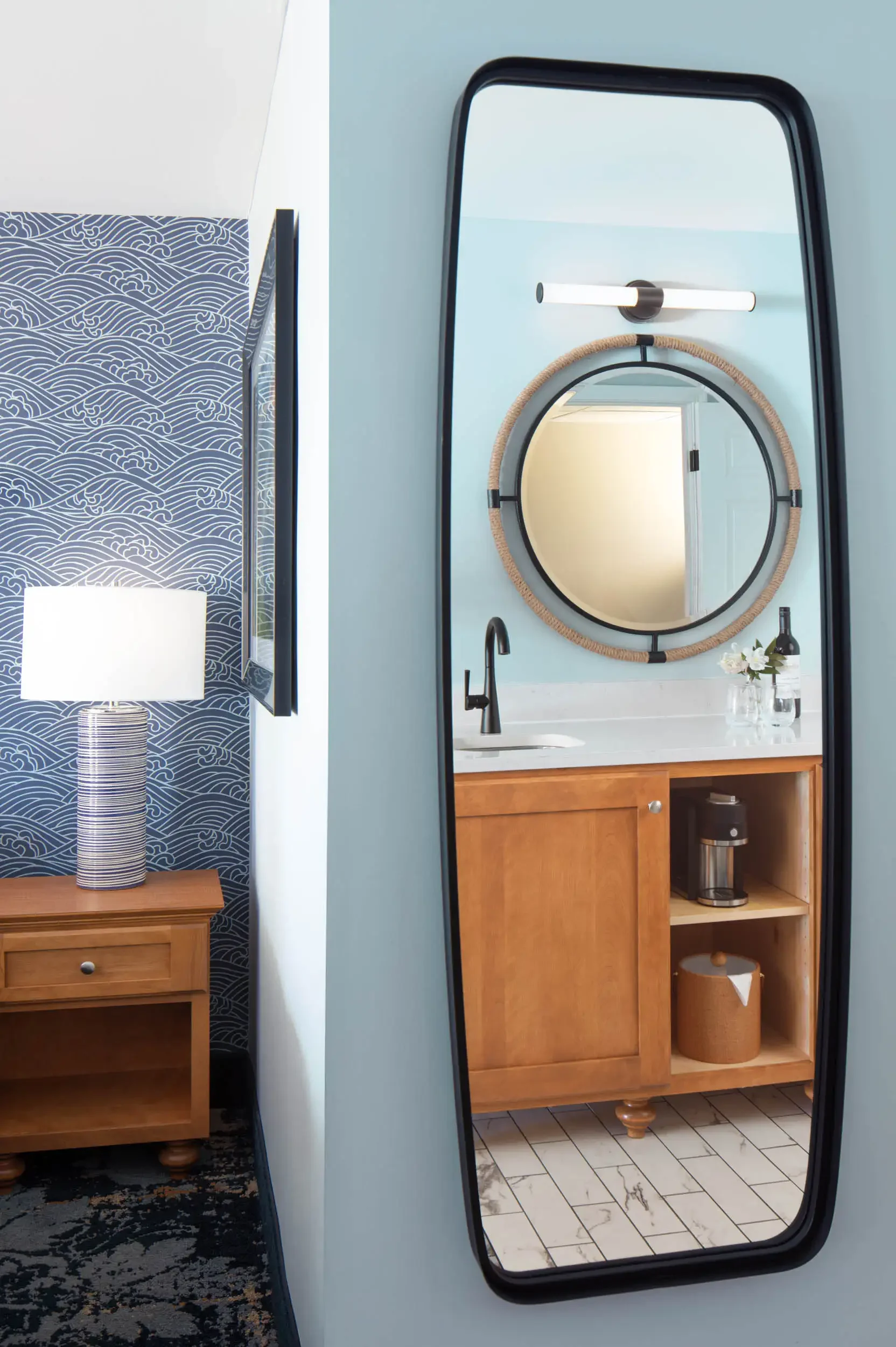 A bathroom with a round mirror framed in rope, a black faucet, wooden vanity, and a small vase with white flowers. The mirror also reflects a door and parts of the ceiling. The floor is tiled in white with black veins.