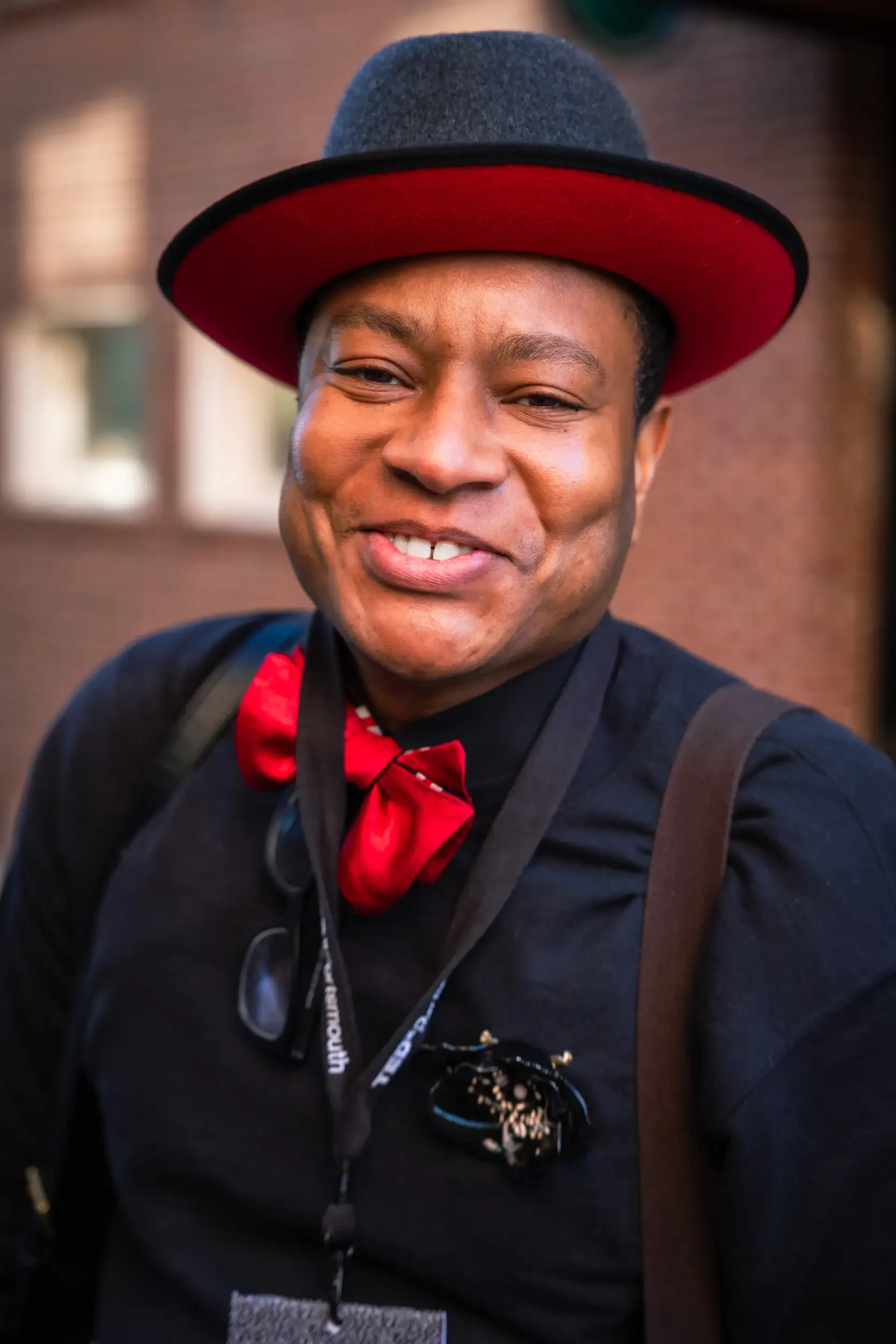A smiling person wearing a gray felt fedora hat with a red underside. They are dressed in a black jacket with a red bow tie, and have motionless sunglasses hanging from their neck. They are outdoors with a blurred background.