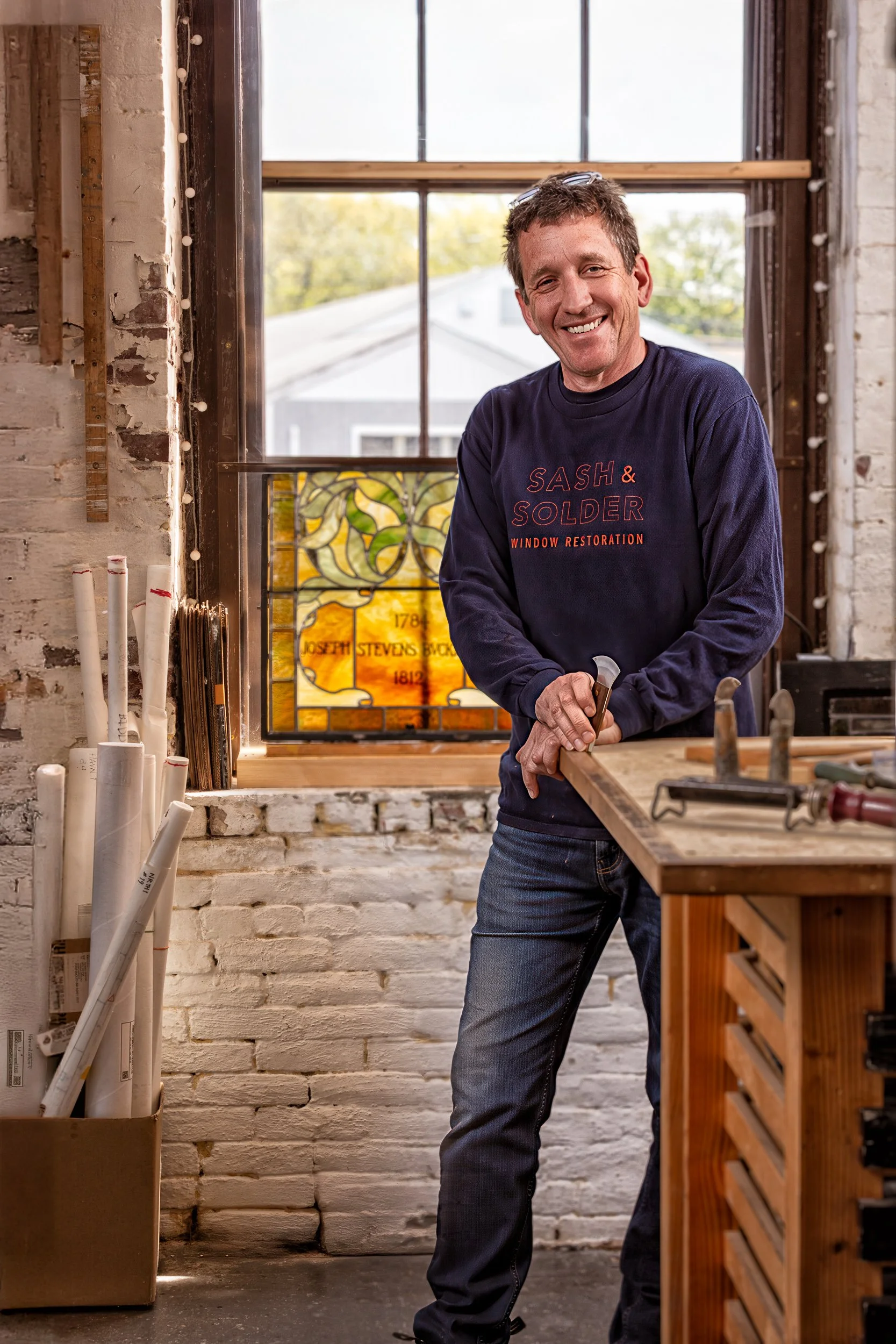 A man smiling inside a workshop, wearing a navy sweatshirt with red text, standing next to a workbench with carpentry tools, in front of a window with a stained glass panel behind him.