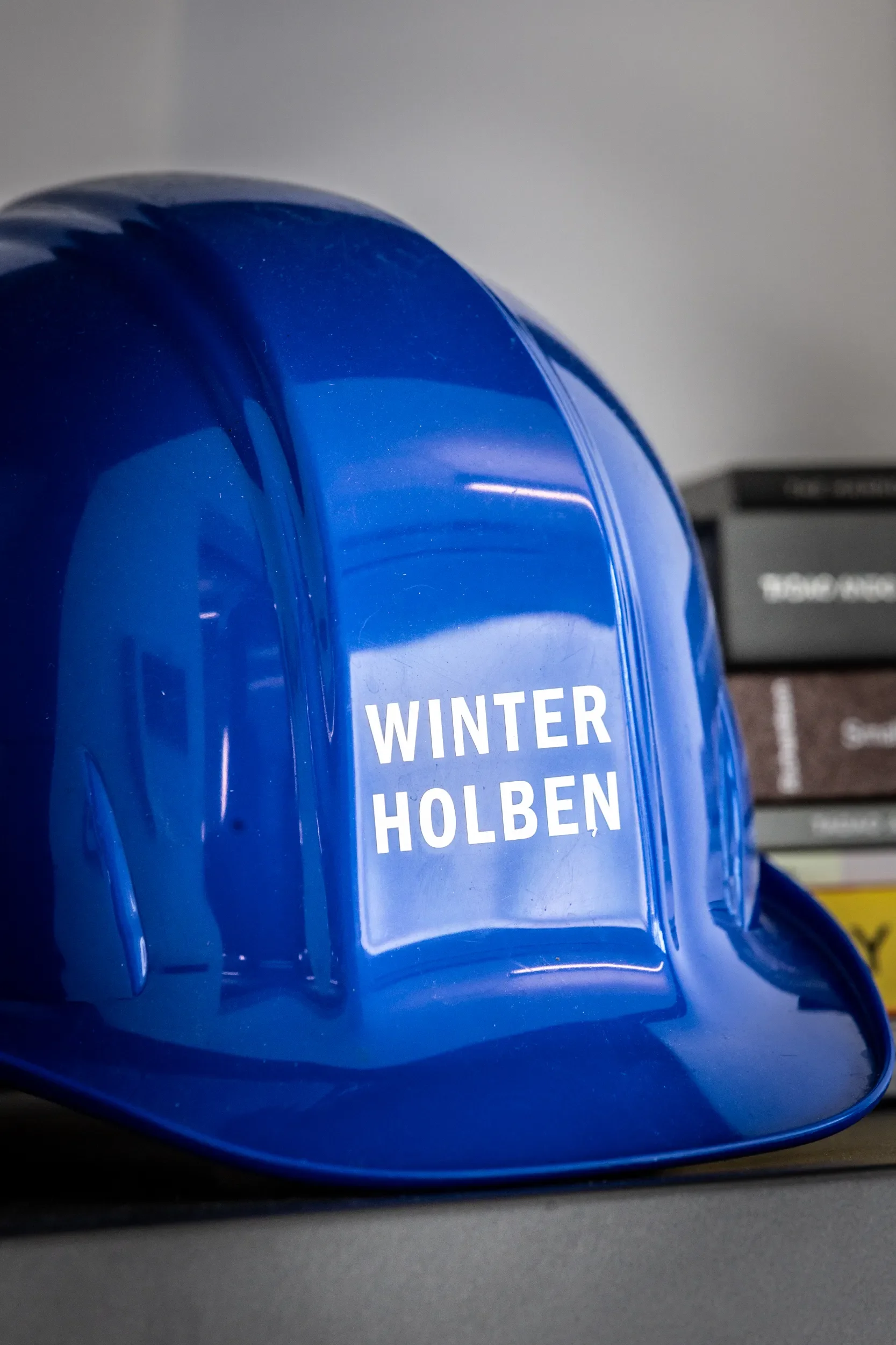 A blue hard hat with the words 'Winter Worker Holben' printed on it, placed on a table in front of a gray background.