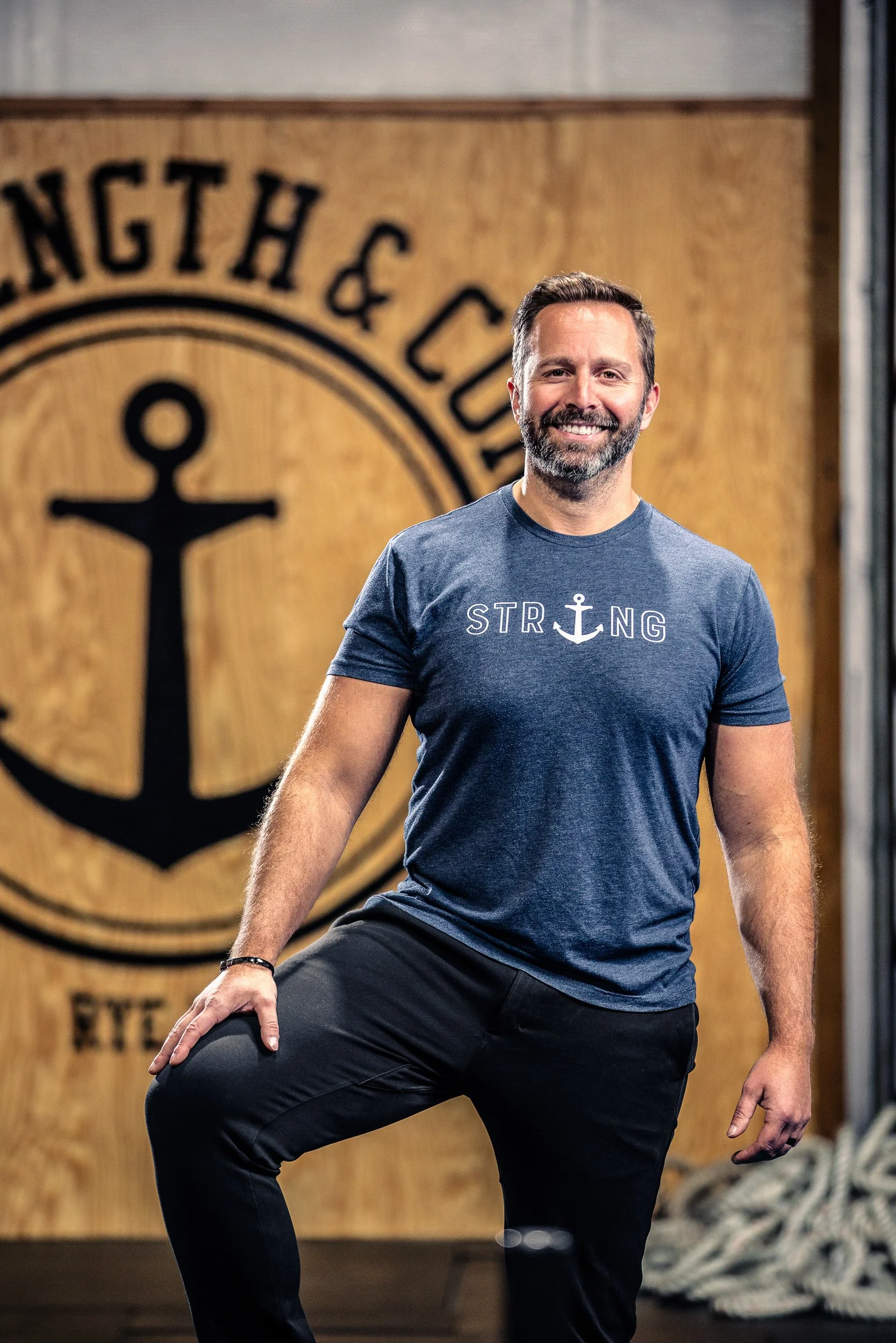 A man with a beard and short hair smiling while standing with one knee raised inside a gym or fitness center. He wears a blue T-shirt with the word 'STRONG' and an anchor symbol on it and dark pants. In the background, there is a wooden wall with a l