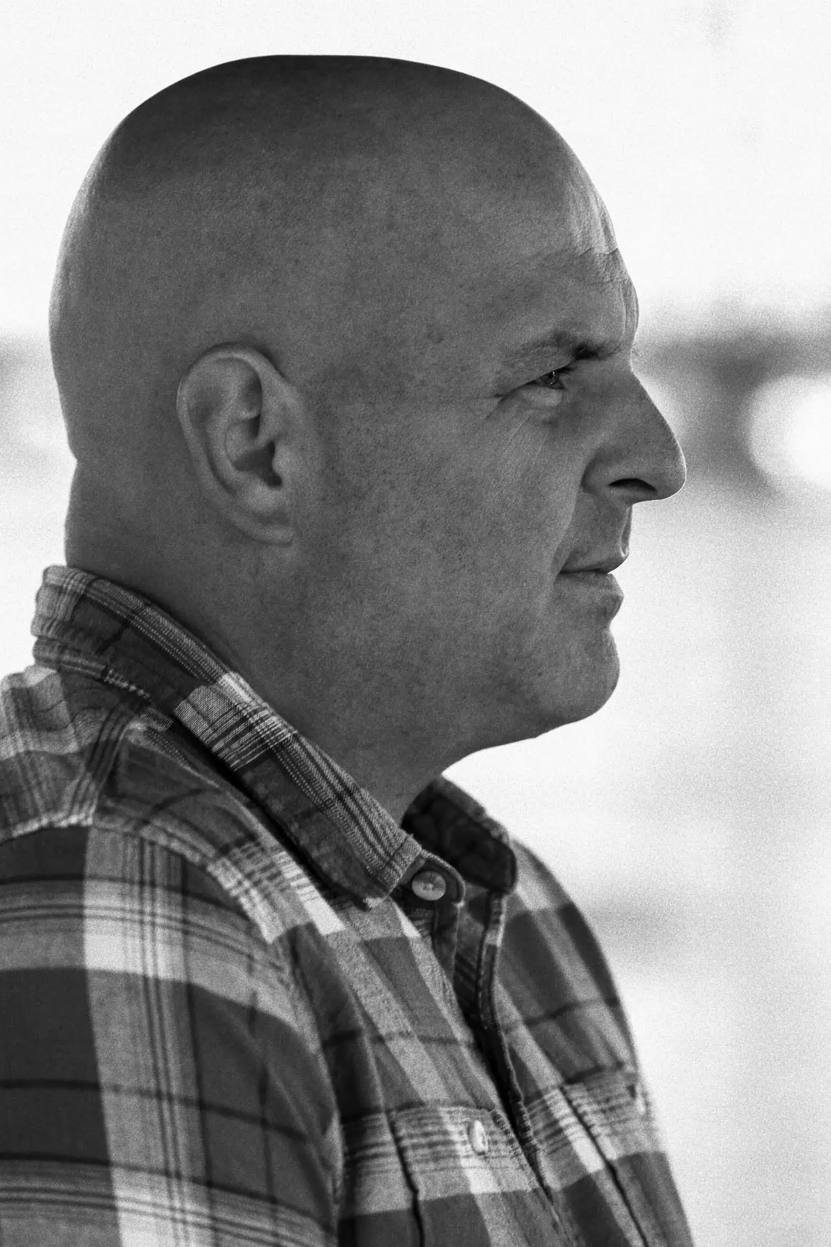 Close-up of a person's side profile with a shaved head, wearing a checkered shirt, looking to the right, in black and white.