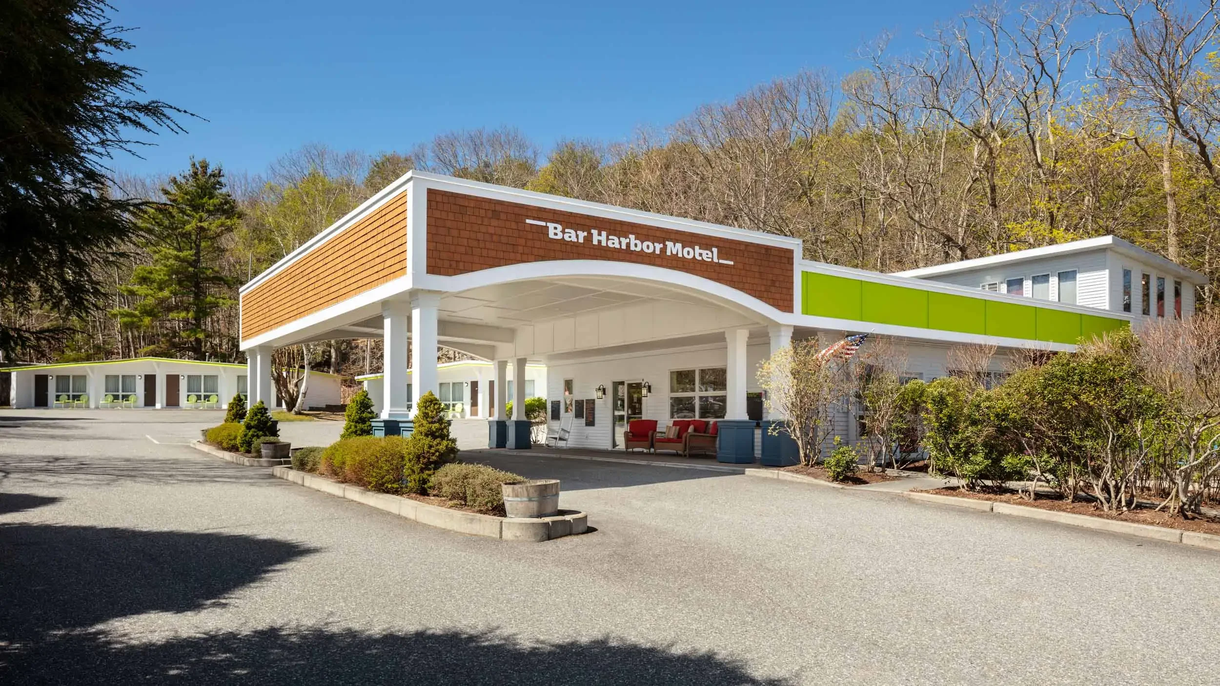Exterior view of the Bar Harbor Motel with a large parking lot, a building with a covered entrance, and a secondary building surrounded by trees, under a clear blue sky.