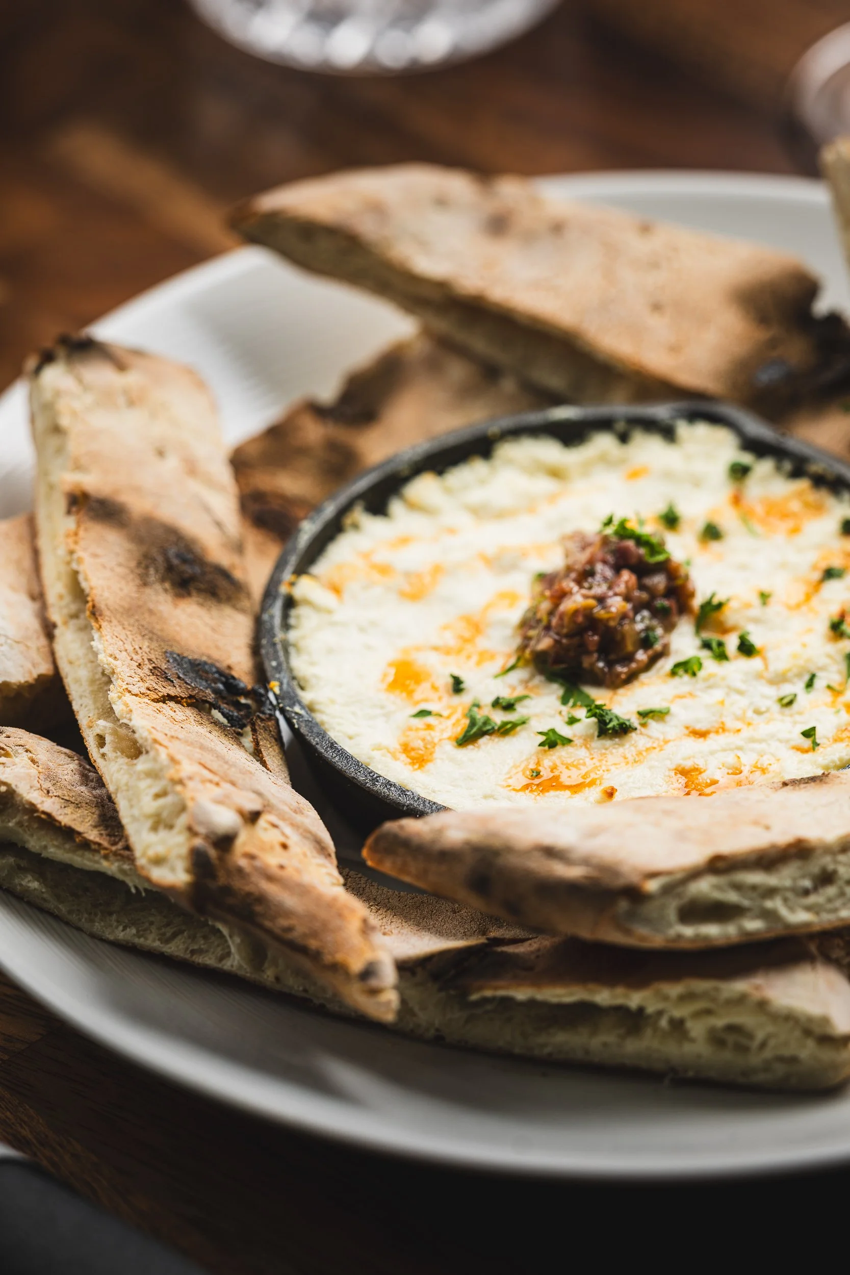A plate of creamy queso dip topped with melted cheese, garnished with herbs, and served with toasted bread slices on a wooden table.