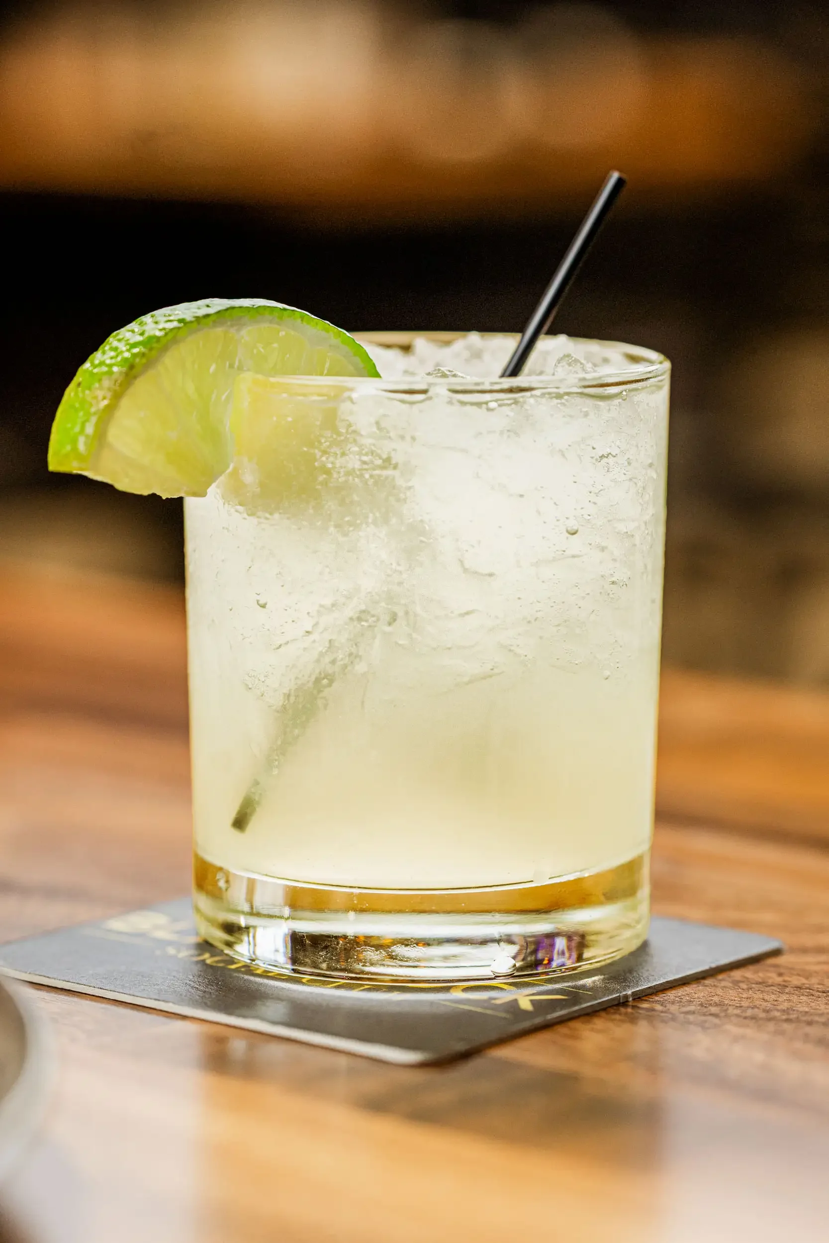 A refreshing cocktail garnished with a lime wedge and served in a glass with ice, placed on a coaster on a wooden table.