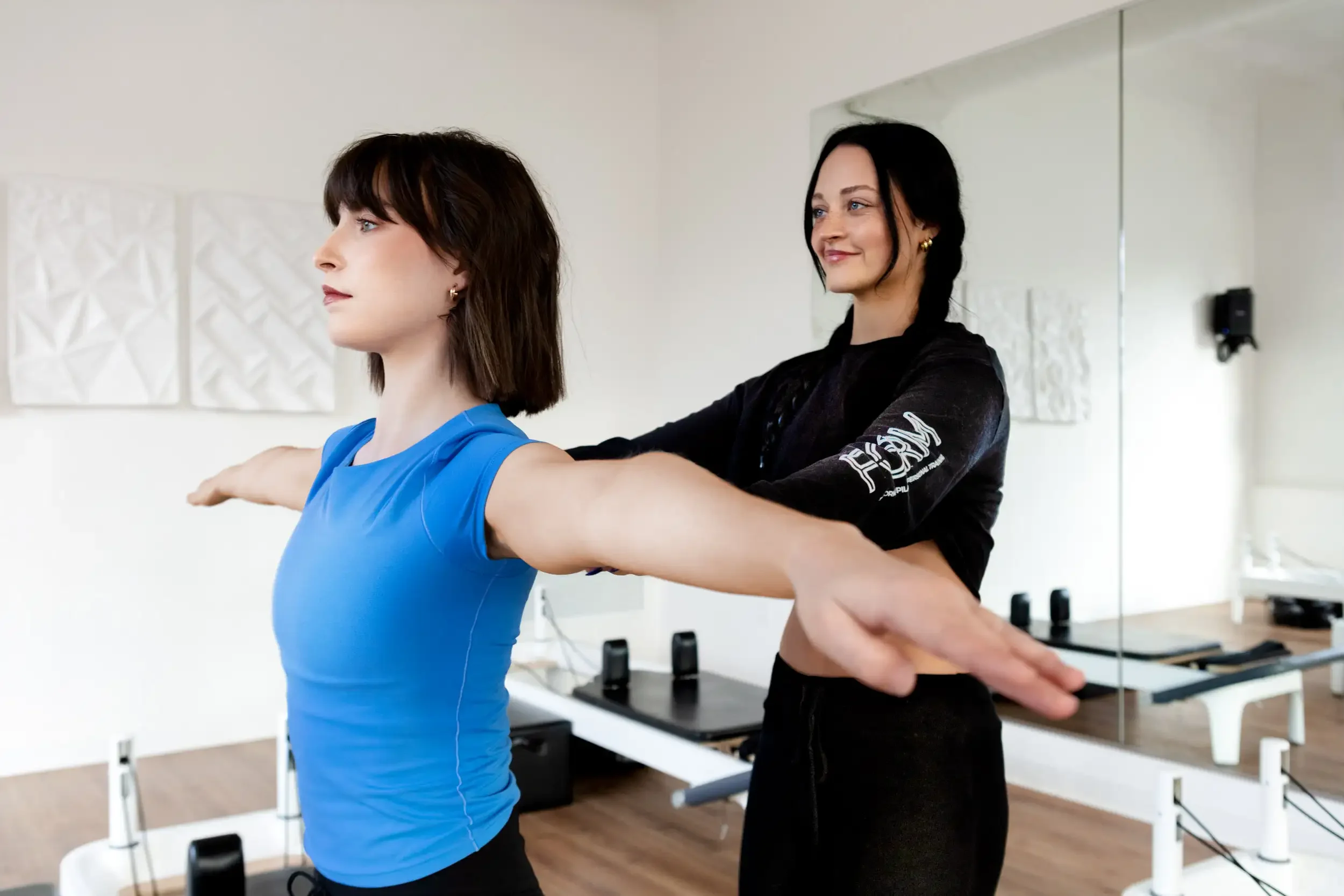 Two women in a fitness studio practicing yoga or stretching exercises, one with dark brown hair and the other with black hair, both facing sideways with arms extended in front of them.