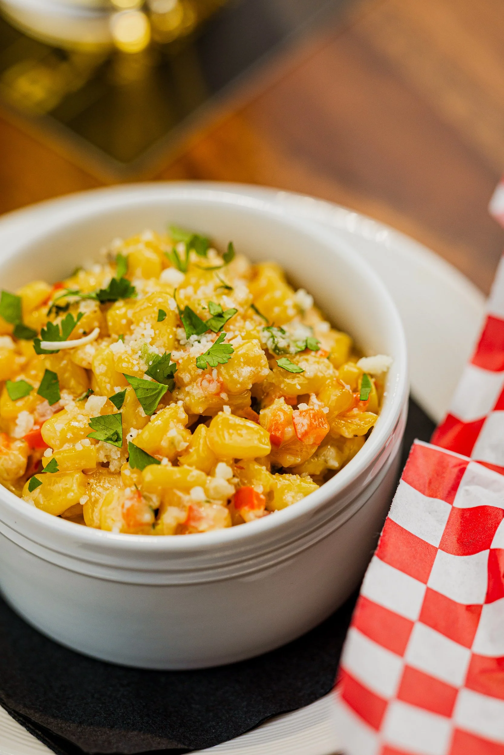 A bowl of corn salad garnished with chopped cilantro, served on a black napkin with a red and white checkered paper around the side of the bowl.
