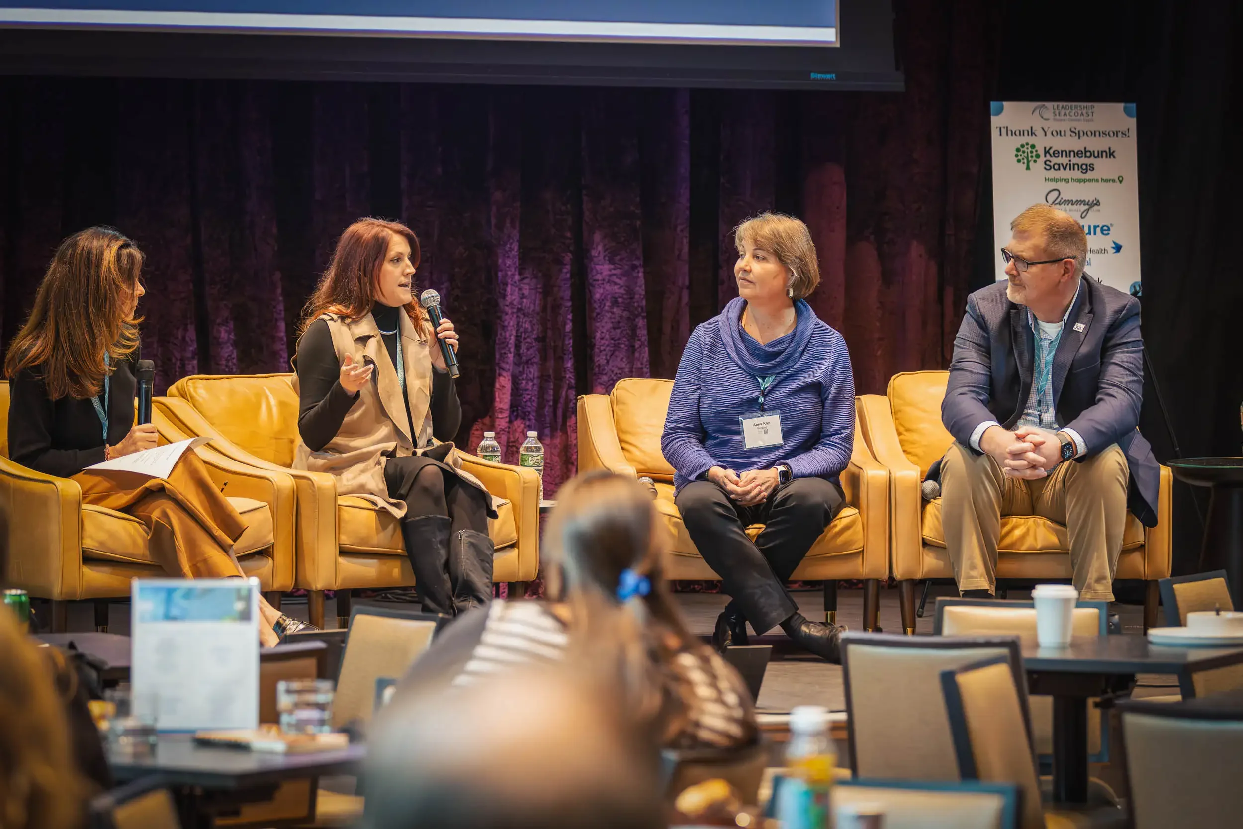 Four people seated on stage, engaged in discussion at a conference or panel. One woman is speaking, holding a microphone, while the others listen. There are water bottles and note papers on the table in front of them, and a banner for sponsors in the