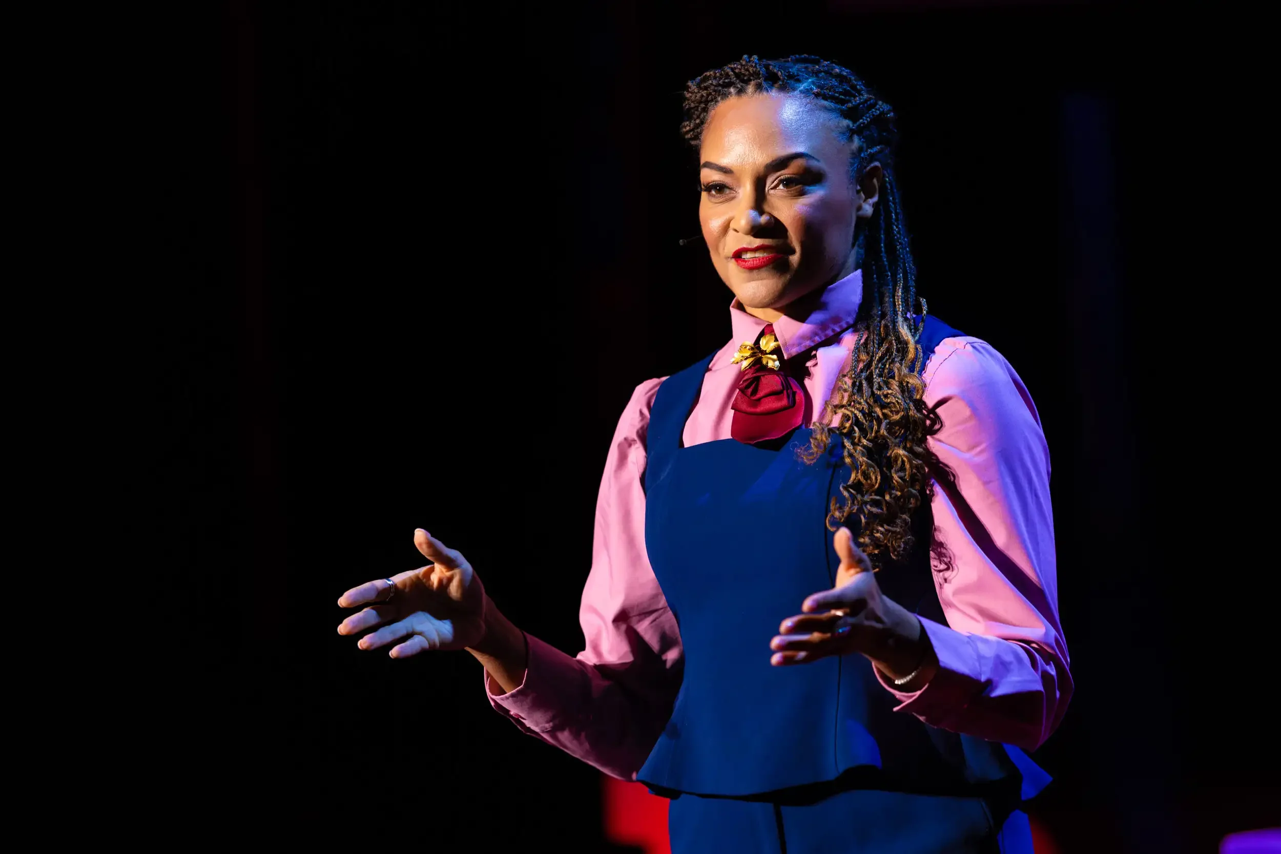 A woman with braided hair giving a presentation on stage, wearing a pink blouse with a bow tie and a navy vest, with a dark background.