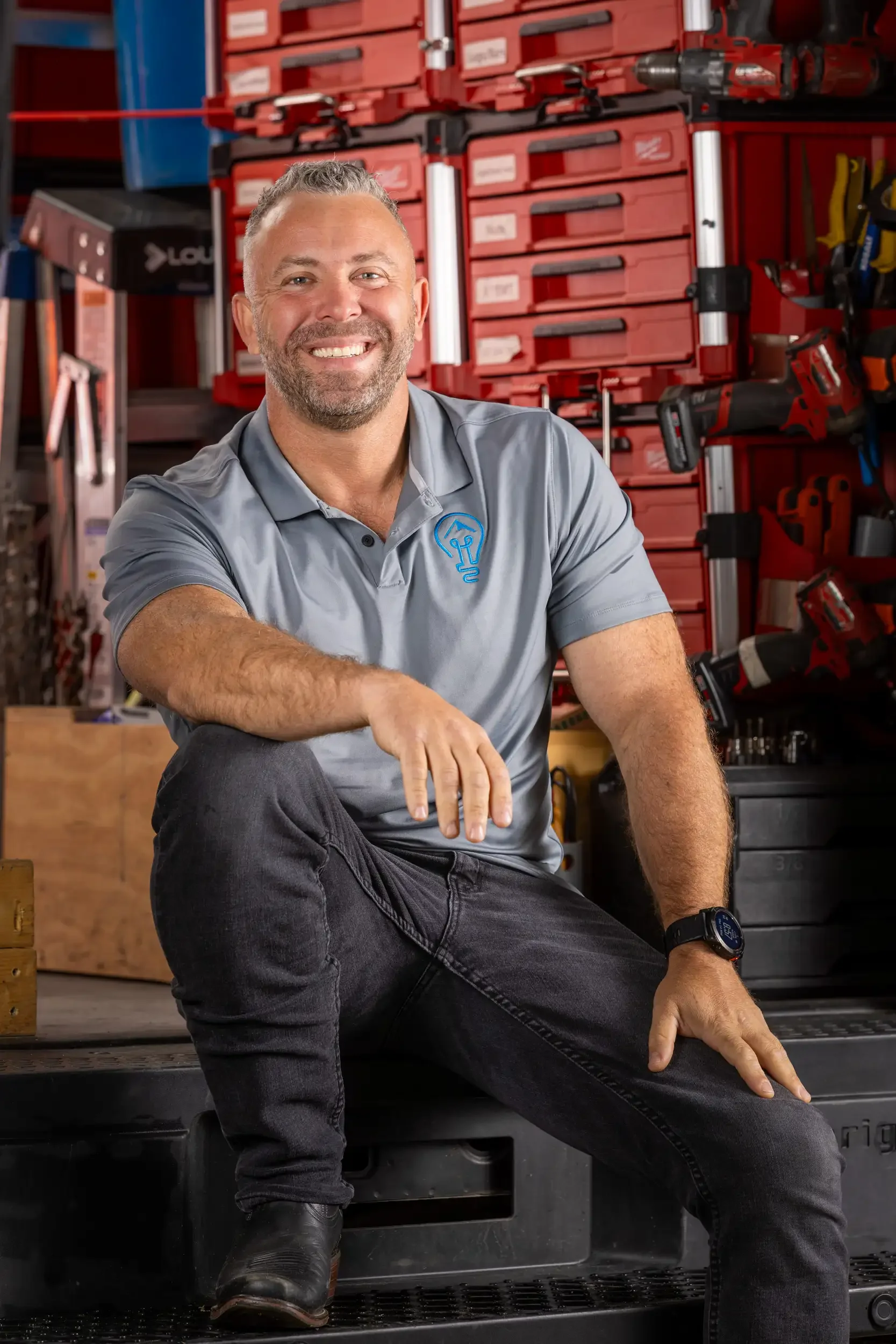 Man smiling and sitting in a workshop surrounded by red storage bins, power tools, and equipment.