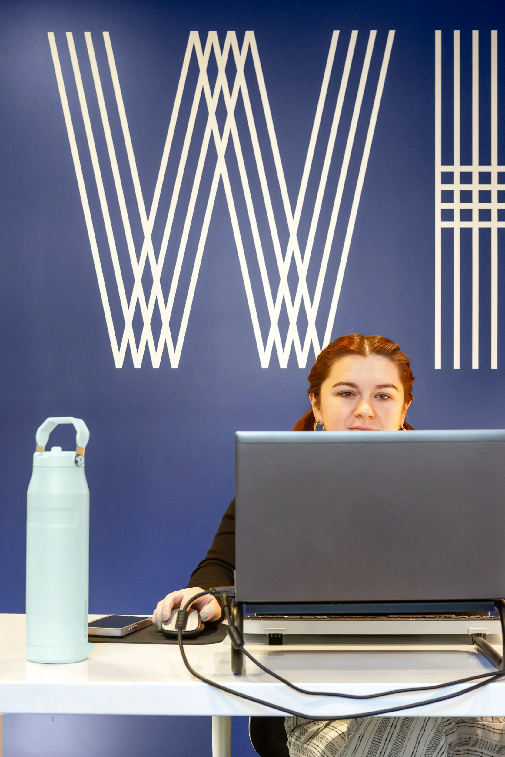 A woman with reddish hair sitting at a desk with a laptop, mouse, and water bottle. She is looking at the camera in front of a blue wall with white geometric design and part of a person's head is visible at the bottom of the image.