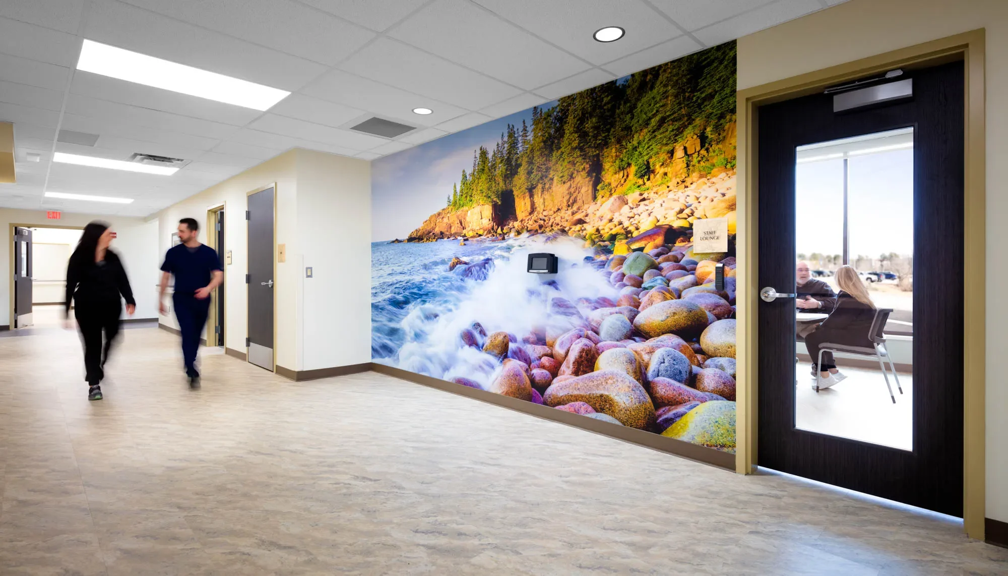 An interior view of a hallway in a building with a large mural of a rocky seashore with a forested coastline on the wall. There are two people walking and talking in the hallway, and a glass door with a sign labeled 'Staff Lounge' shows two people sitting at a table inside.