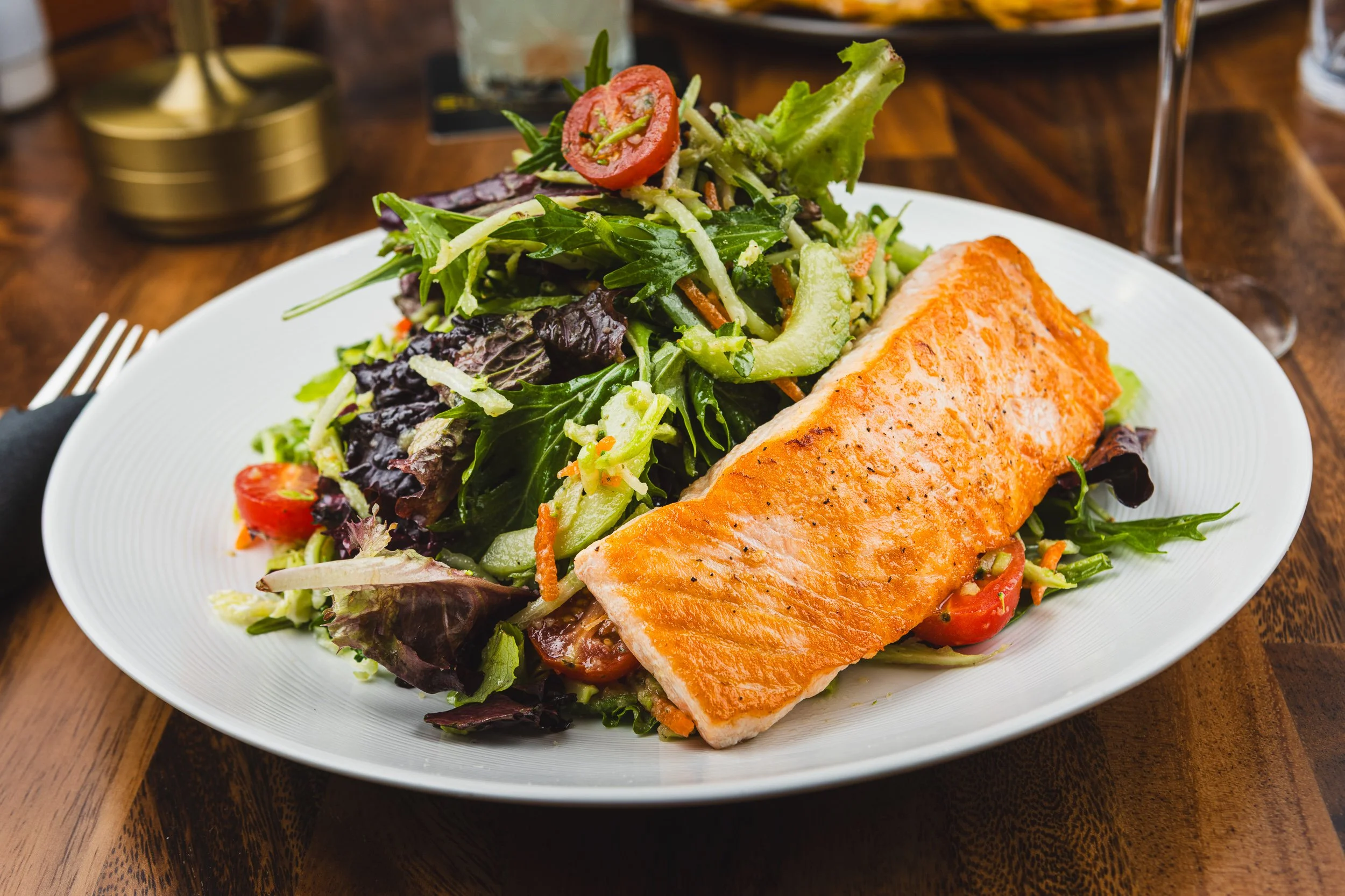 Grilled salmon fillet served with mixed green salad including lettuce, cherry tomatoes, cucumbers, and sliced vegetables on a white plate.