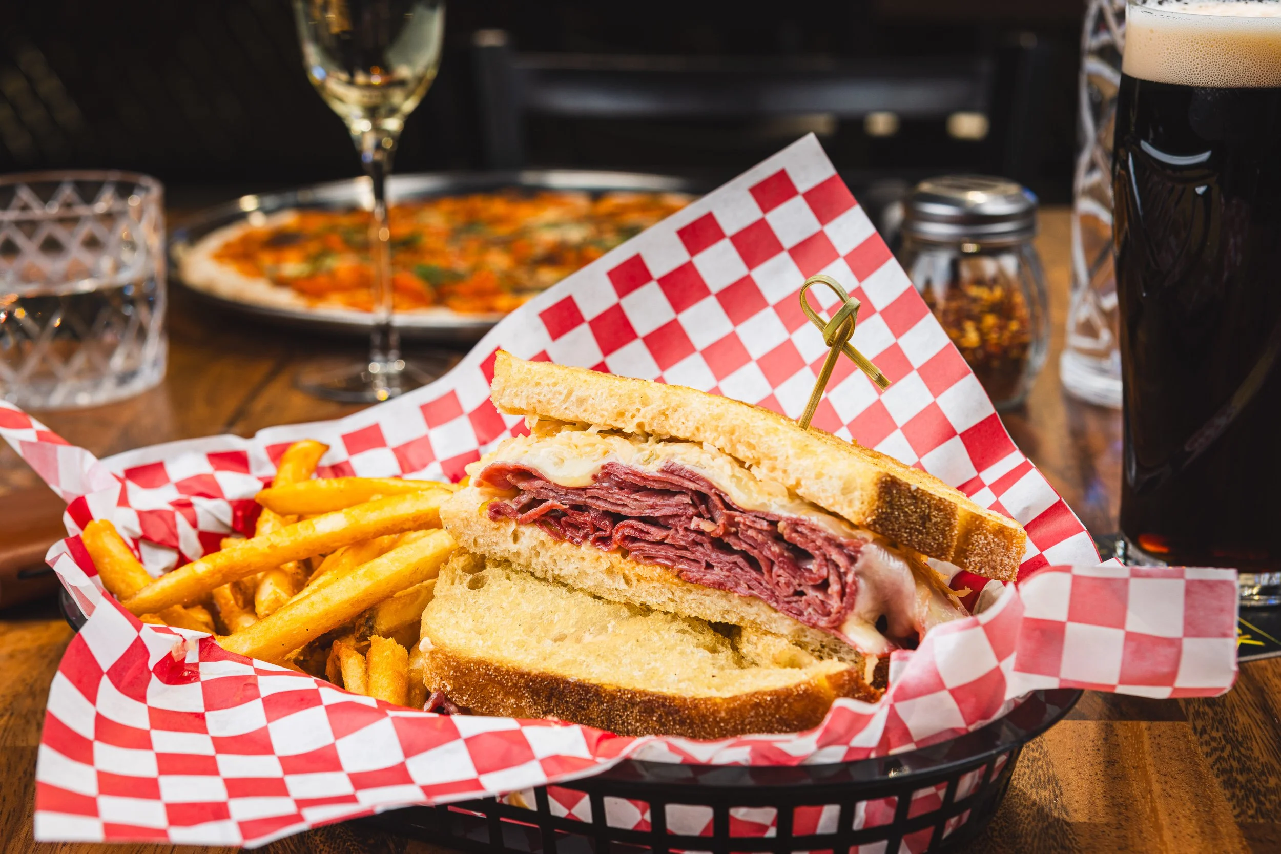 A cheeseburger with sliced roast beef and melted cheese, served with French fries in a red and white checkered basket on a wooden table, with drinks and pizza in the background.